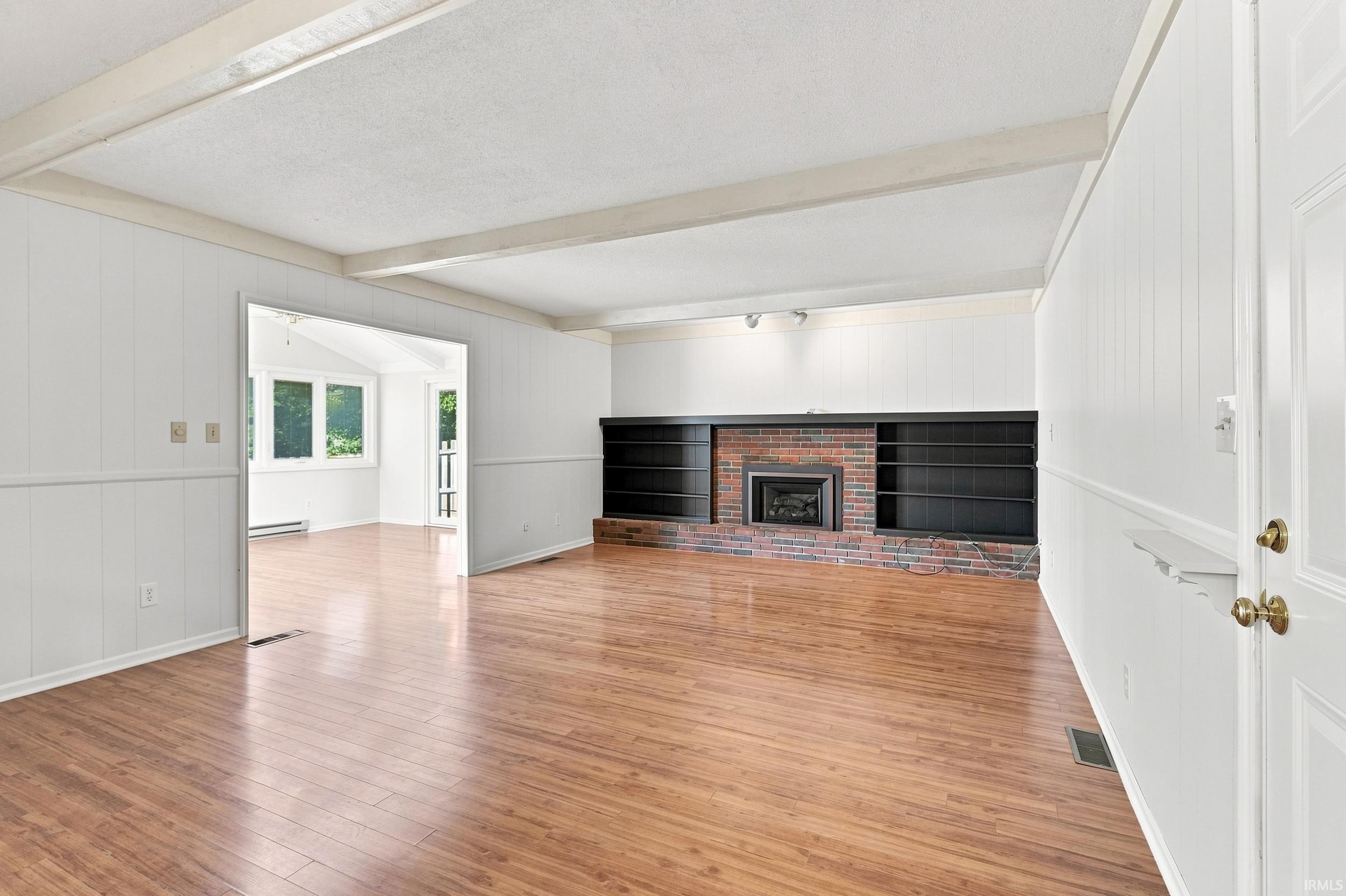 Spacious open-concept living area featuring gleaming wood-finish flooring, soaring coffered ceilings with recessed lighting, and a striking brick fireplace with black surround and hearth