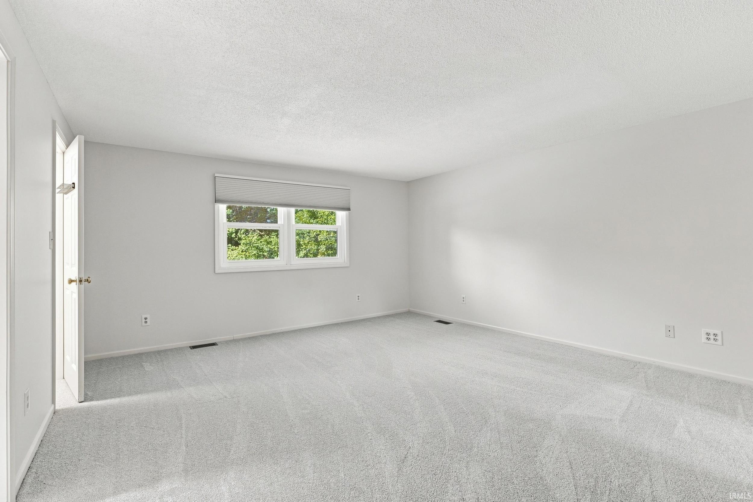 Spacious room featuring light gray carpeting, neutral wall paint, and a window with a roller shade