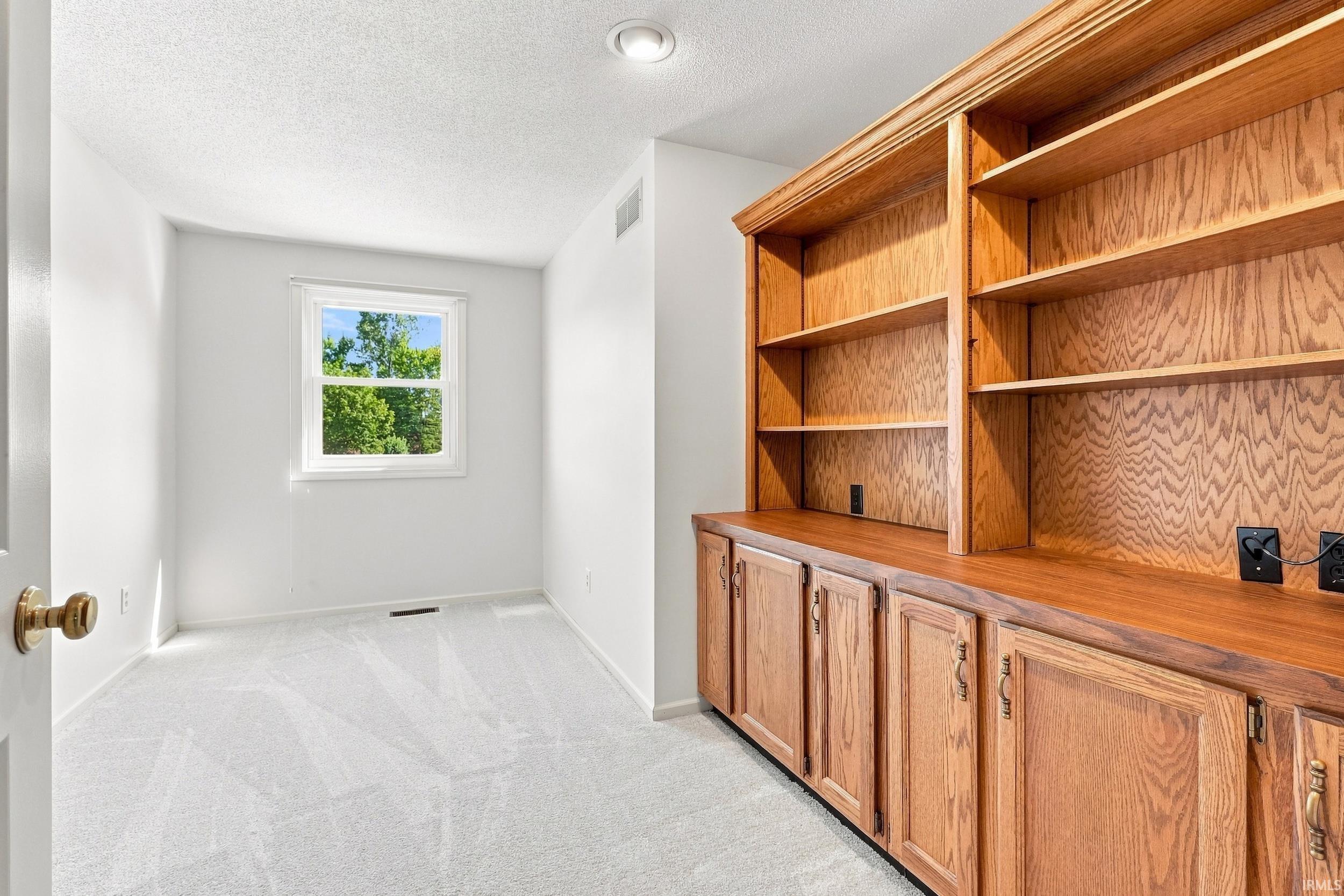Room featuring a large built-in wood cabinetry unit with integrated shelving and base cabinets, recessed ceiling lighting, a window with exterior tree scenery, and light-colored patterned carpeting