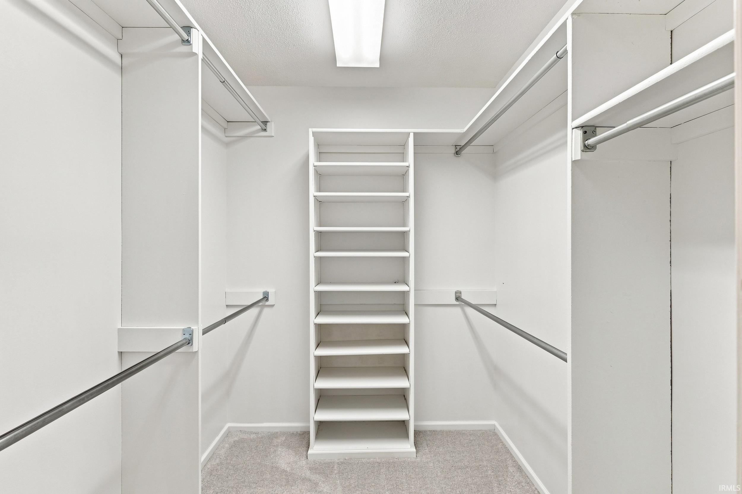 Spacious walk-in closet featuring floor-to-ceiling white shelving with multiple storage levels, chrome hanging rods on both sides, recessed ceiling lighting, and neutral carpeting