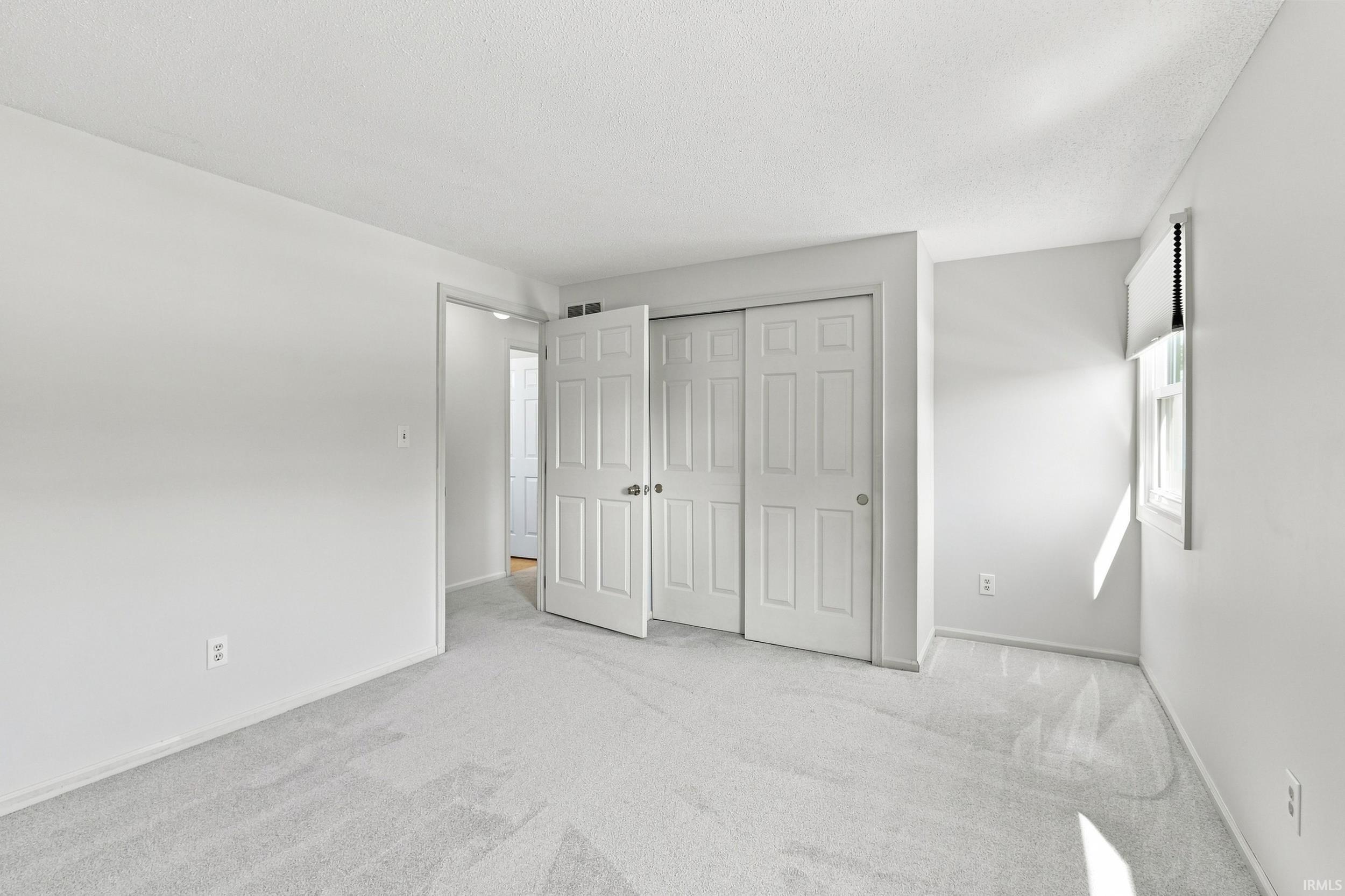 Light gray wall paint, plush carpet flooring, a window with roller blinds, and a closet with bifold and sliding doors