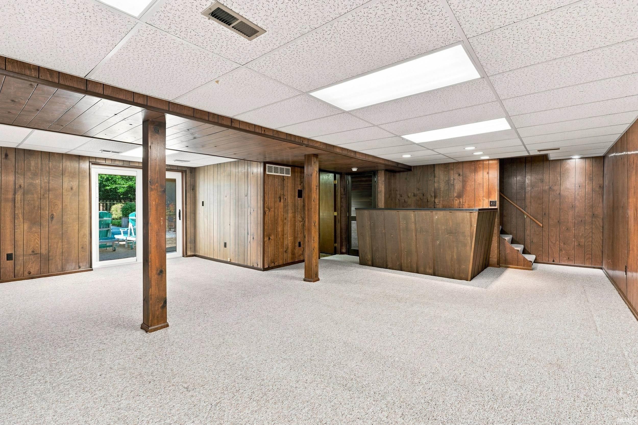 Lower-level finished space featuring wood-panel walls, light-colored carpet, recessed lighting, and a built-in wet bar with a staircase