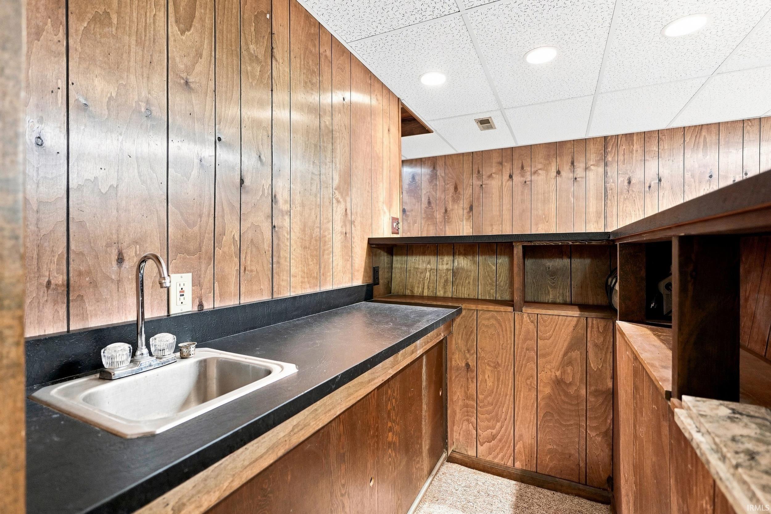 Wet bar area featuring wood paneling, black countertops, a stainless steel sink with a chrome faucet, and recessed ceiling lighting