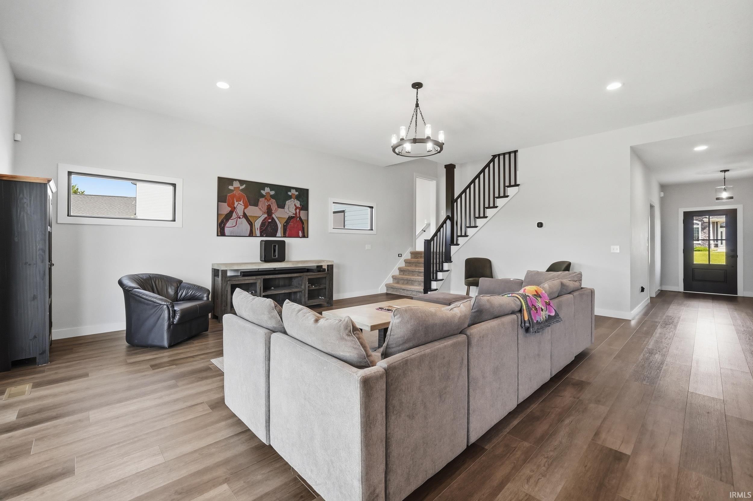 Expansive living area featuring wood-finish flooring, a contemporary chandelier, a staircase with black railings, recessed lighting, and a front entry door with glass panels