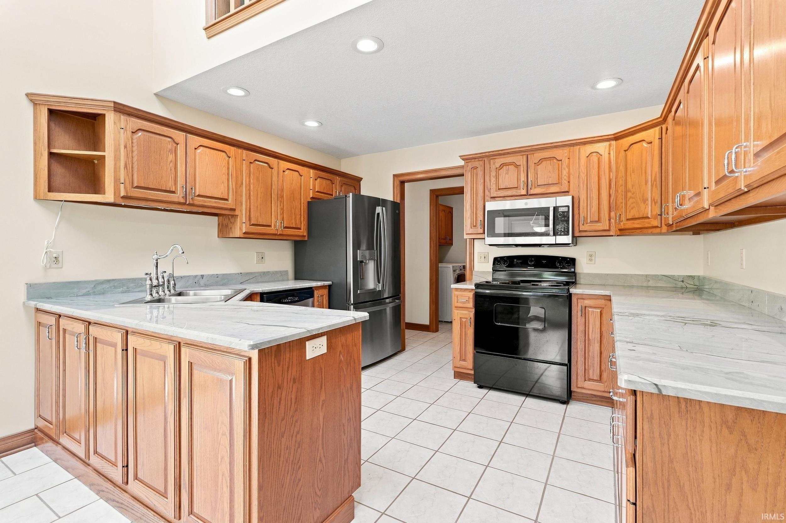 Spacious kitchen featuring warm wood cabinetry throughout, a large center island with countertop seating, stainless steel appliances including a French-door refrigerator and built-in microwave, tile flooring, and recessed ceiling lighting