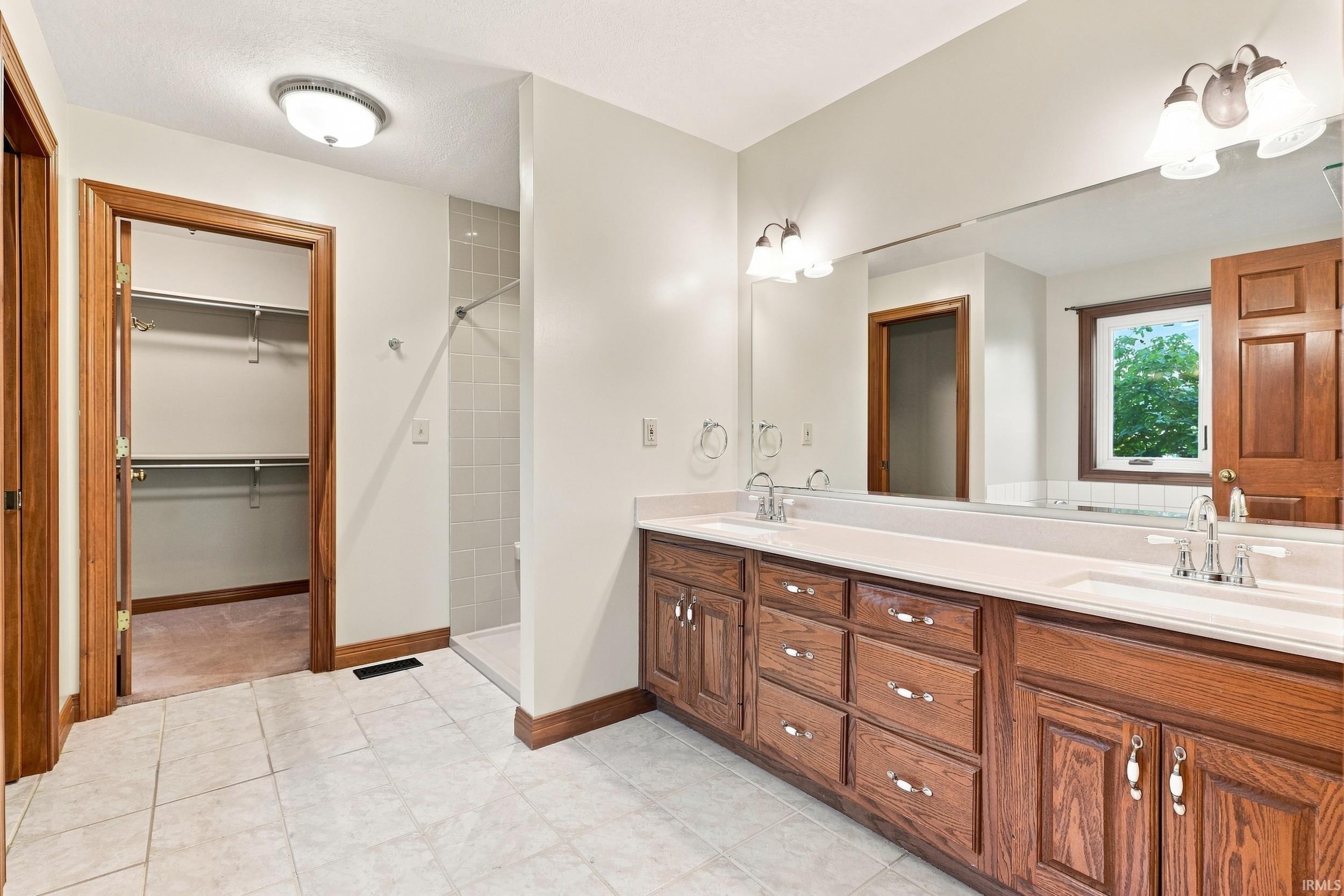Bathroom featuring a double vanity with wood cabinetry and a continuous countertop, a large wall-mounted mirror, ceramic tile flooring, and a tiled shower stall