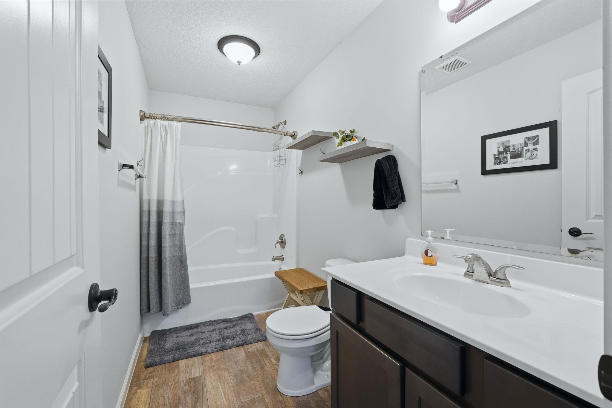 White-paneled door with dark hardware, wood-finish flooring, a white bathtub with a shower kit, a dark wood-finish vanity with an integrated sink, and wall-mounted shelving