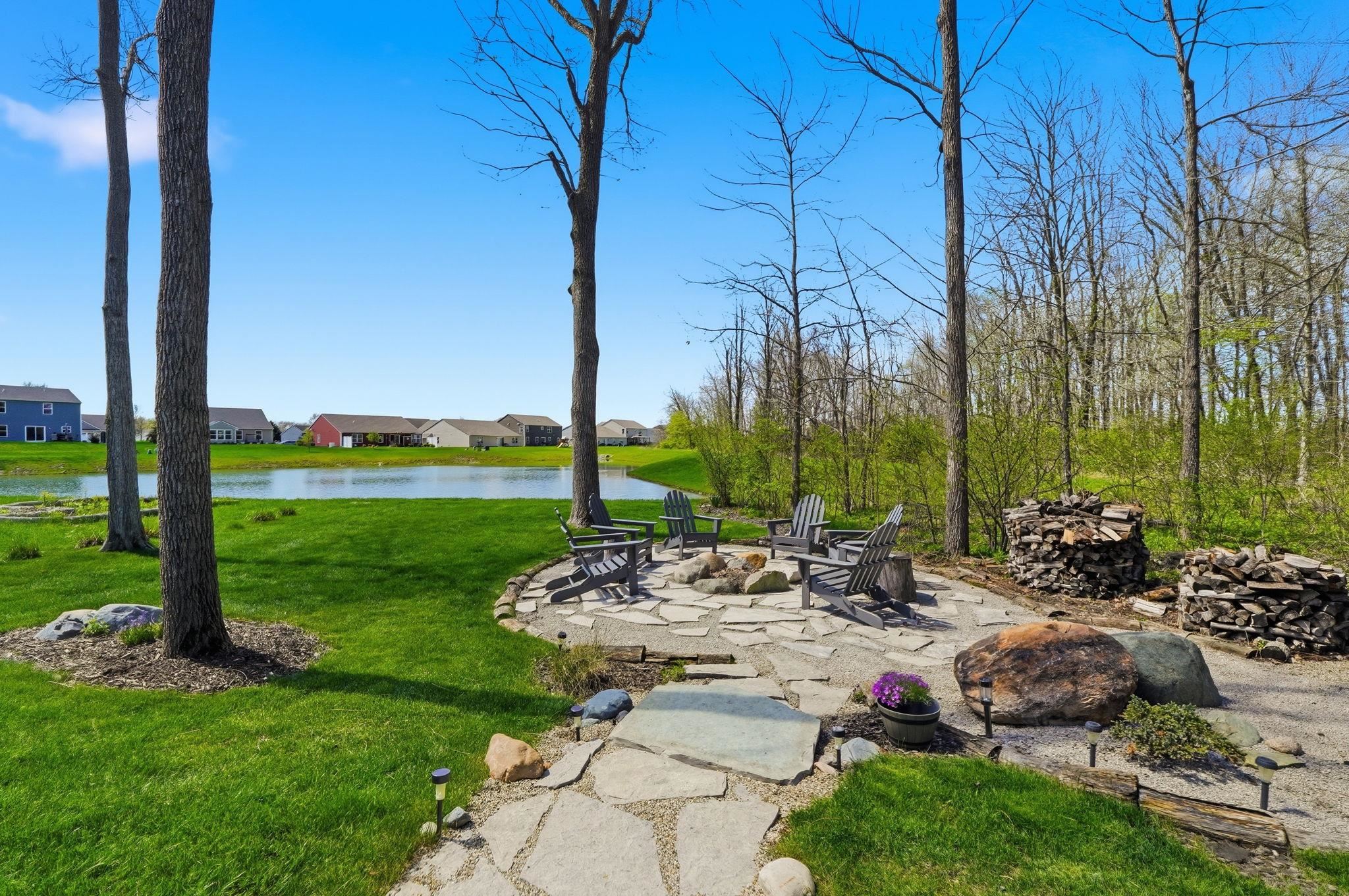 Stone patio with fire pit, surrounded by Adirondack chairs, mature trees, and a pond