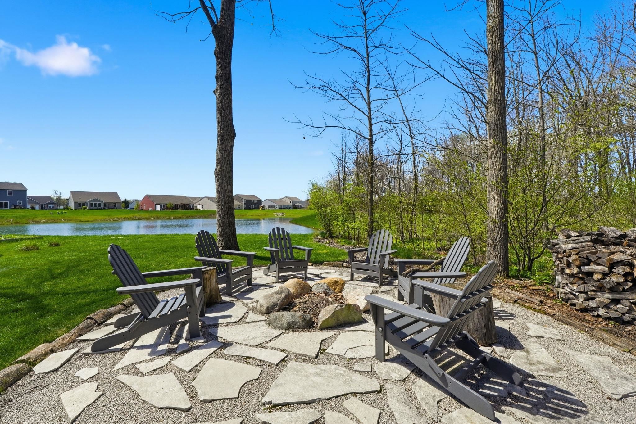 Outdoor fire pit area featuring a stone and gravel patio, surrounded by mature trees, with a water feature in the background