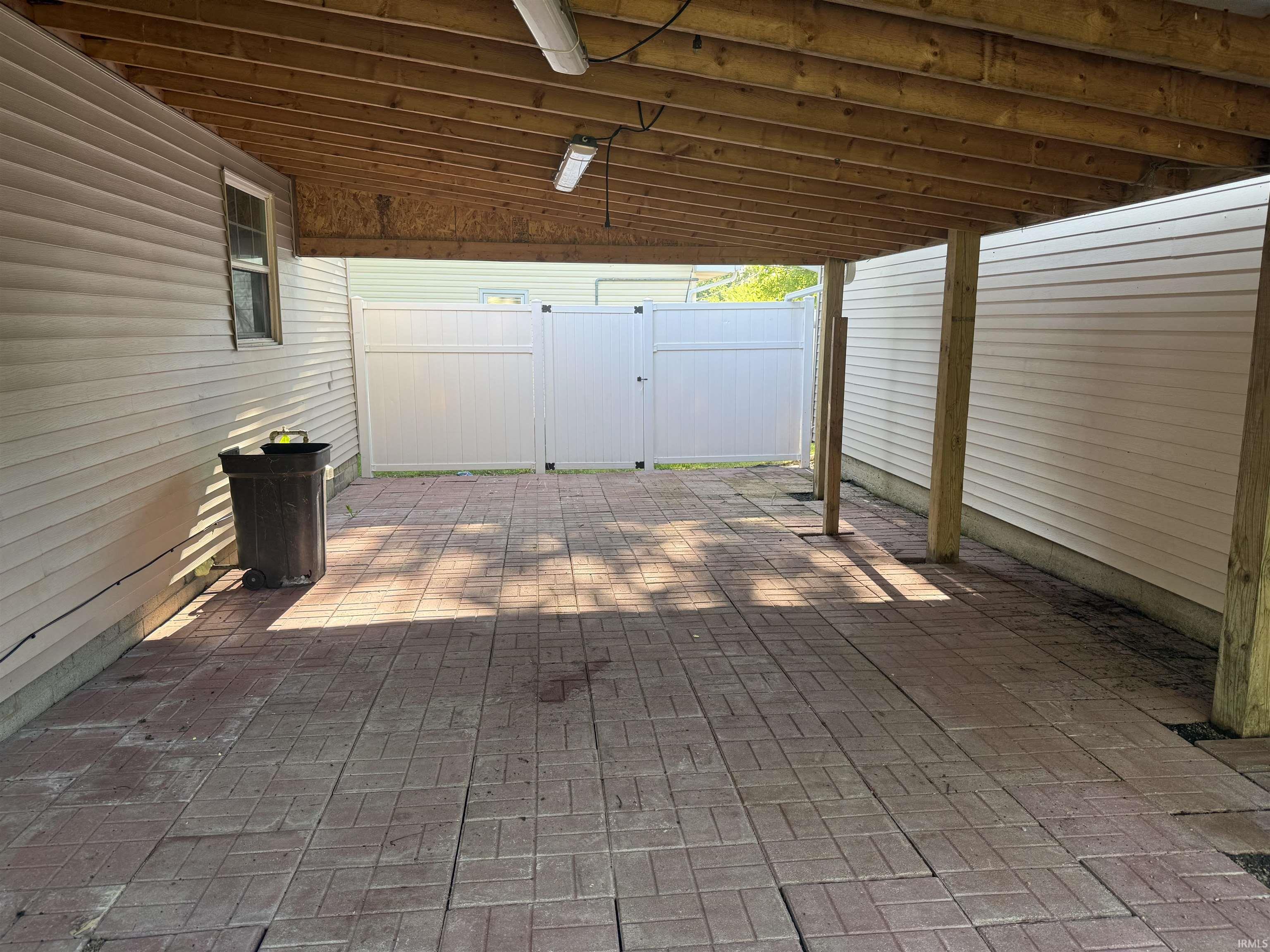 Covered outdoor area featuring a red paver patio, exposed wood beam ceiling, wood support posts, and a white vinyl fence