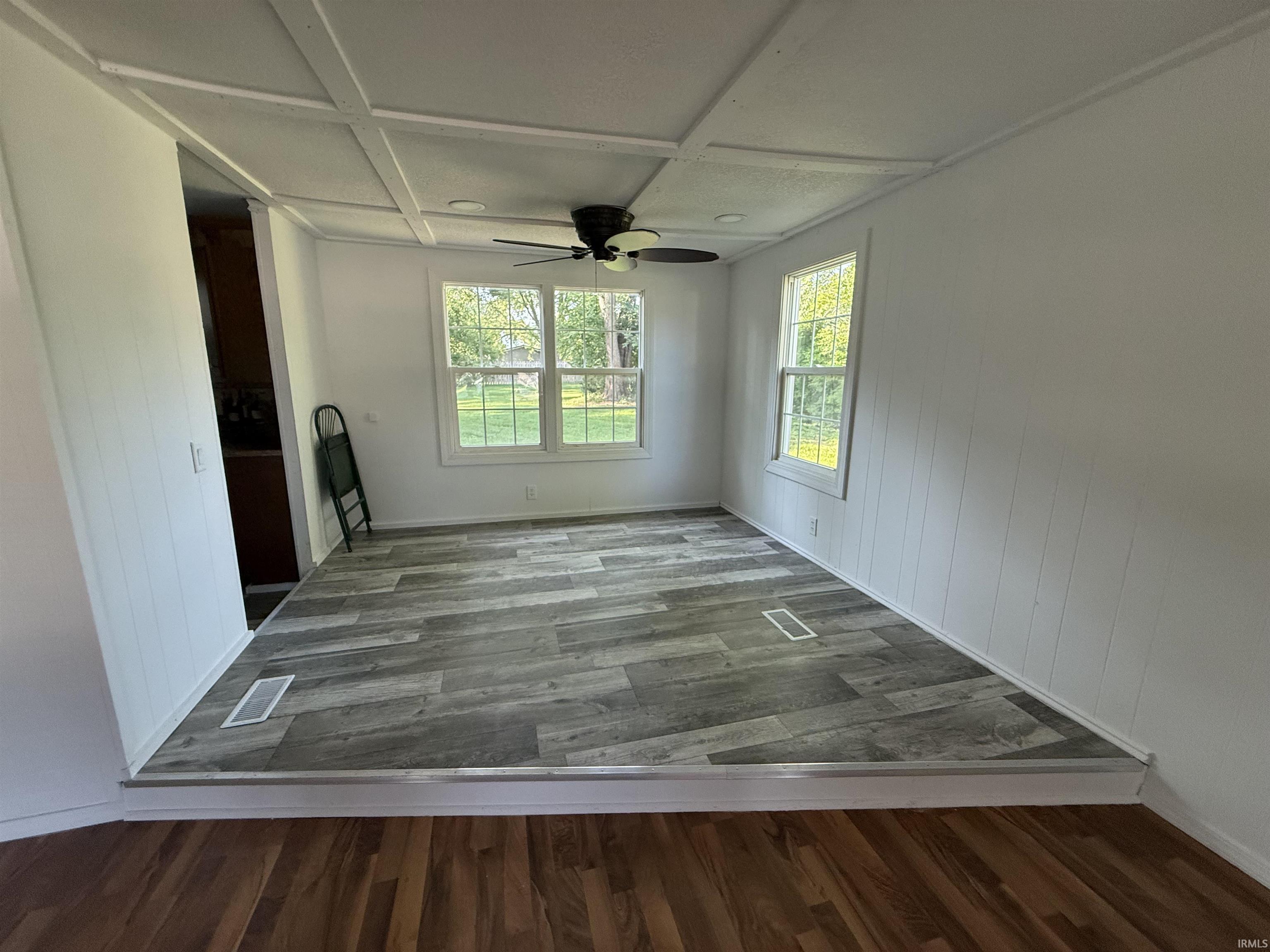 Bright room featuring wood-finish flooring, white vertical wall paneling, a ceiling fan with integrated lighting, and two multi-pane windows