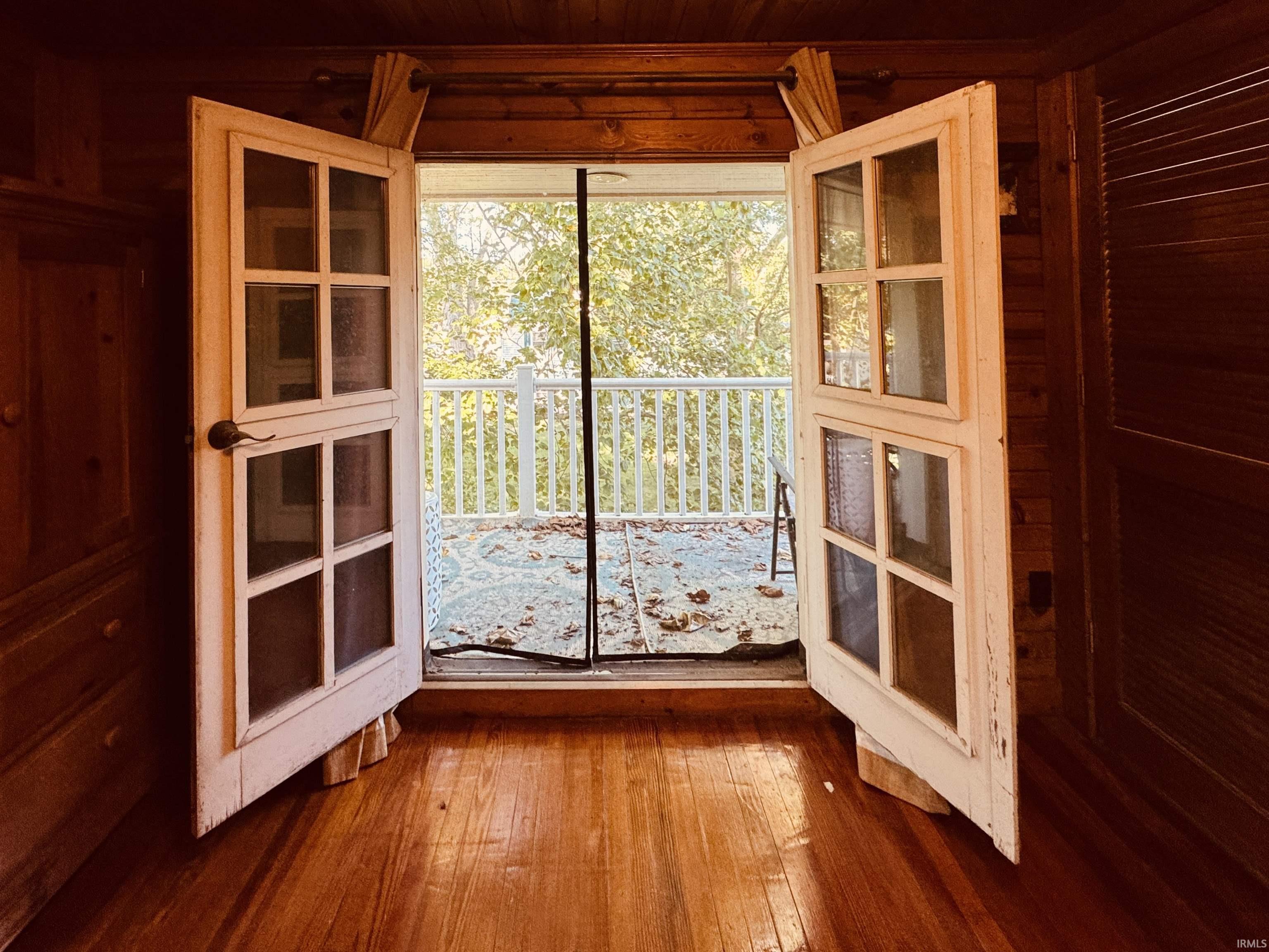 Wood-finish flooring leading to a balcony with white railings, featuring open French doors with glass panes, and wood-paneled walls