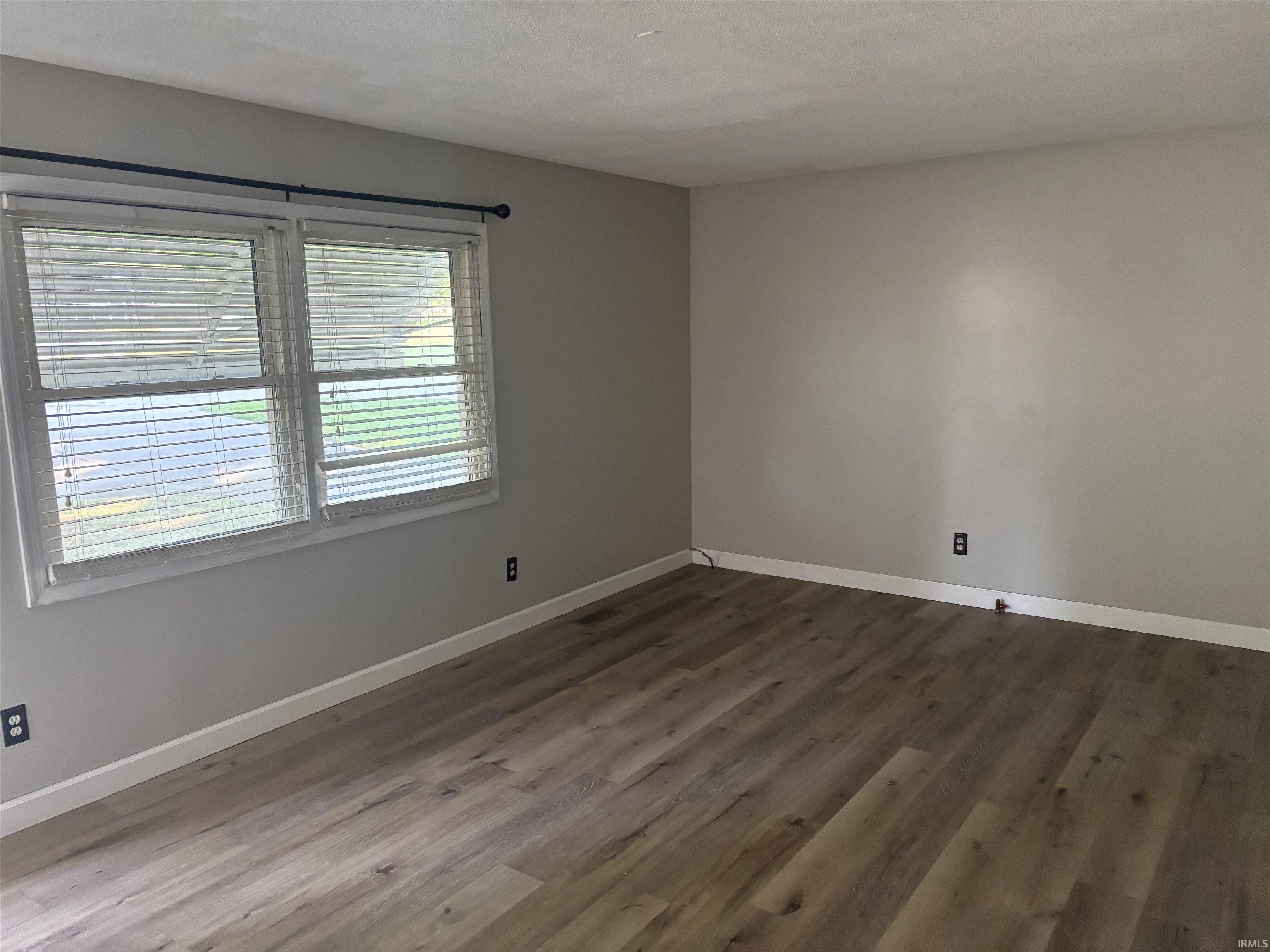 Spacious room featuring wood-finish flooring, light grey painted walls, white baseboards, and a double window with white blinds and a black curtain rod