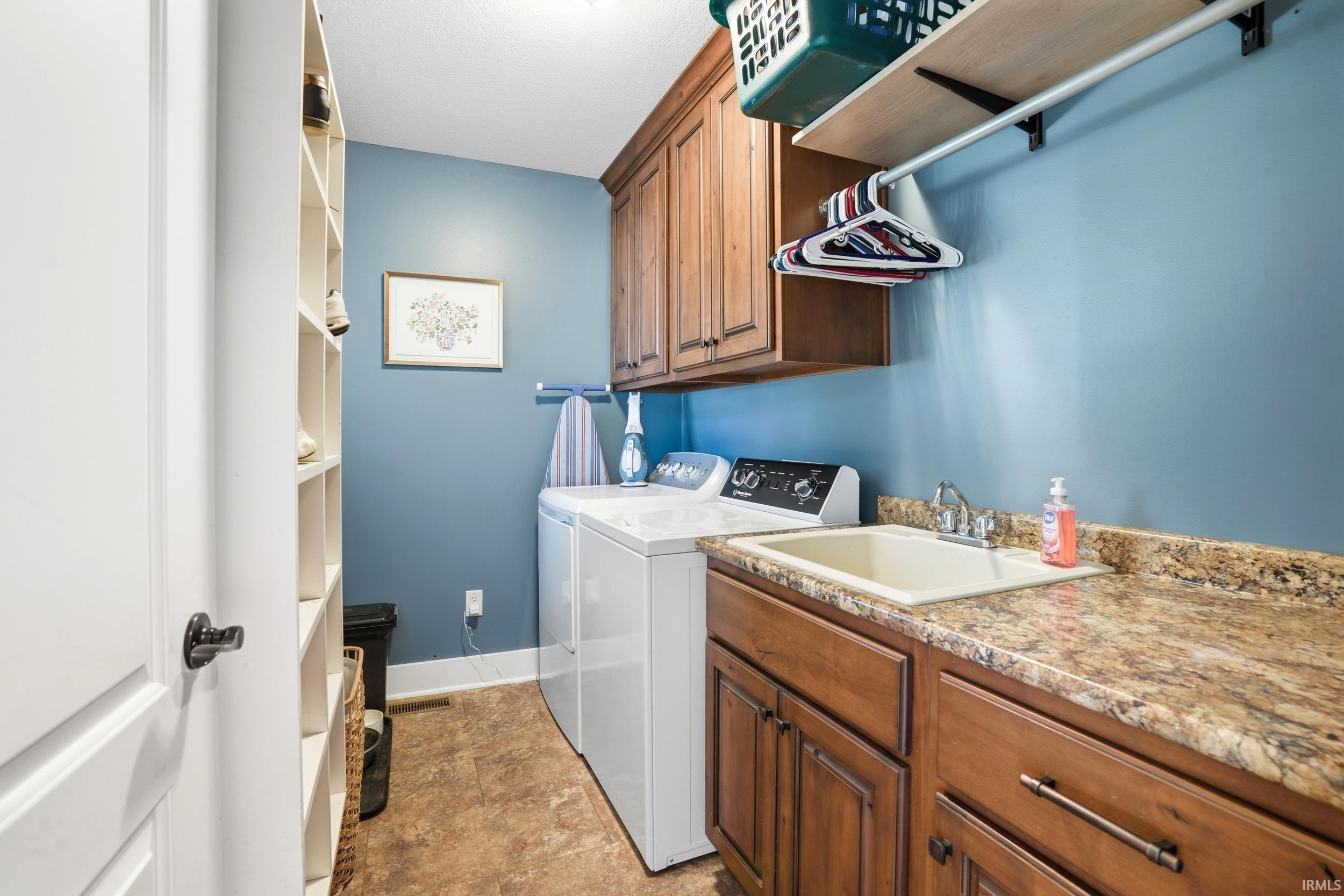 Dedicated laundry room featuring wood-finish cabinetry, granite-style countertops, white front-load washer and dryer, stainless steel sink, abundant storage with wall-mounted shelving, and soft blue accent walls with cork-finish flooring throughout