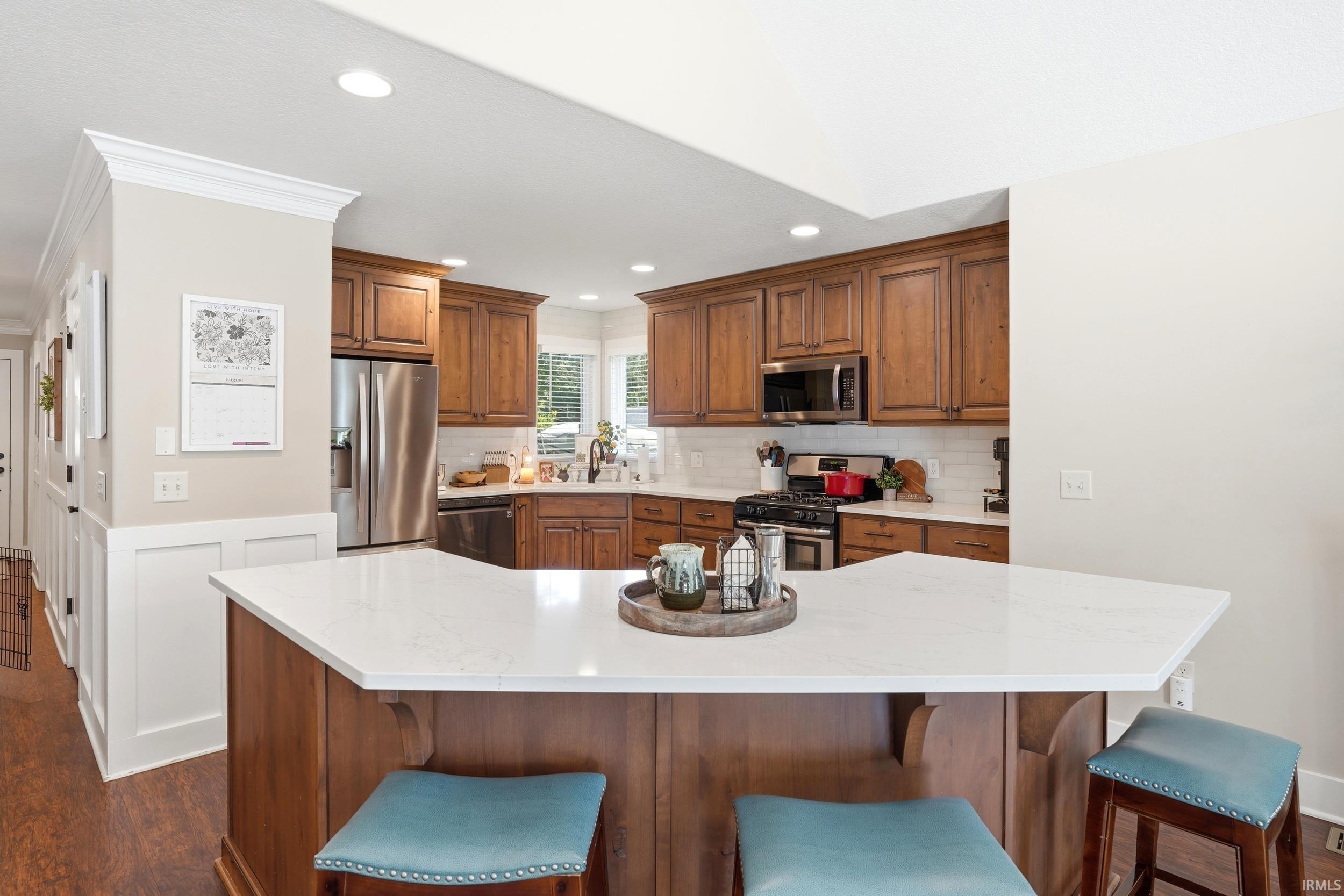 Kitchen featuring an angled island with a light-toned countertop, wood-finish cabinetry, stainless steel appliances, white subway tile backsplash, and recessed lighting