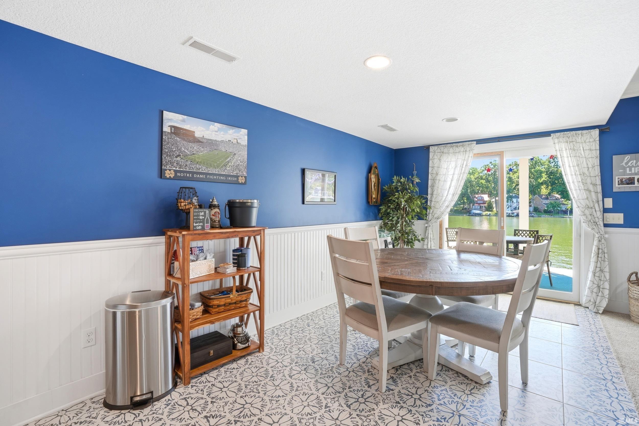 Bright and airy dining area showcasing bold blue accent walls paired with crisp white wainscoting, ceramic tile flooring with geometric patterning, and abundant natural light from French doors and windows overlooking the grounds