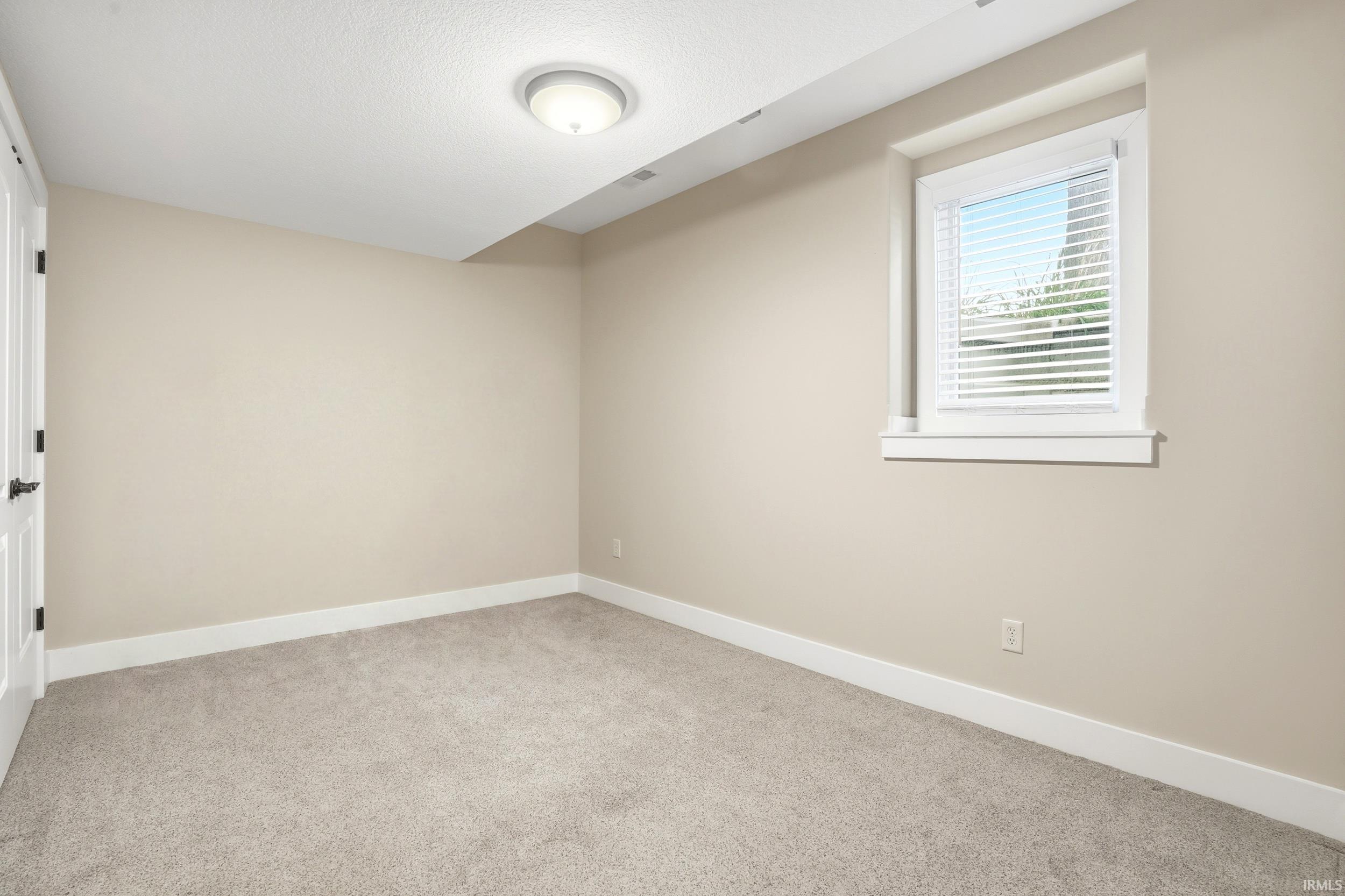 Spacious bedroom featuring neutral beige walls, plush carpet flooring, and a large casement window with integrated blinds providing natural light
