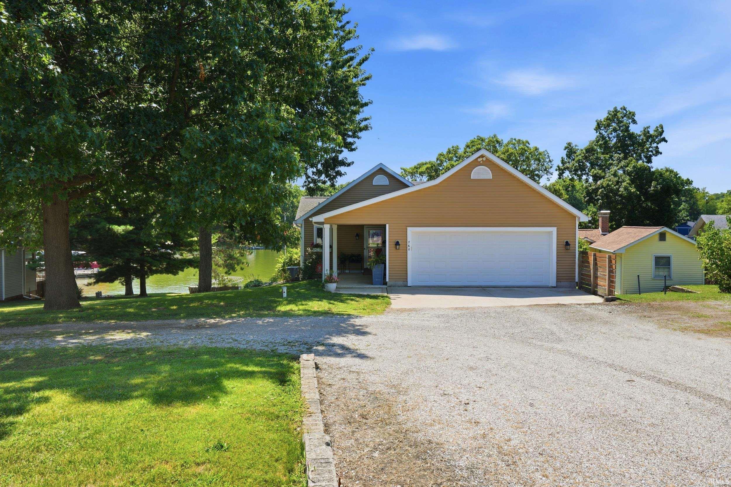 Tan exterior home featuring a two-car garage with white paneled door, situated on a level lot with mature shade trees and established landscaping