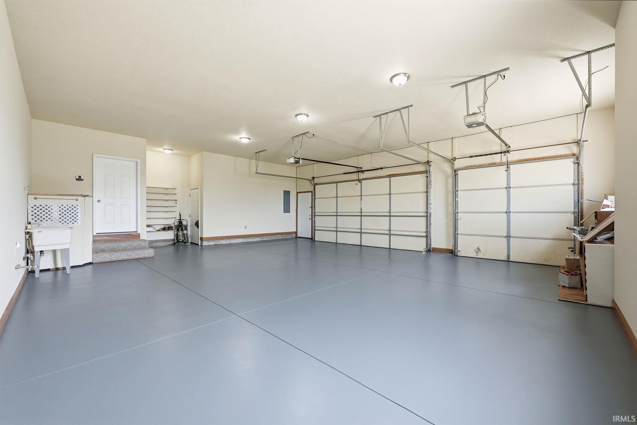 Three-car garage featuring a gray epoxy-coated floor, two garage door openers, multiple flush-mount ceiling lights, a utility sink, and interior access via a white door