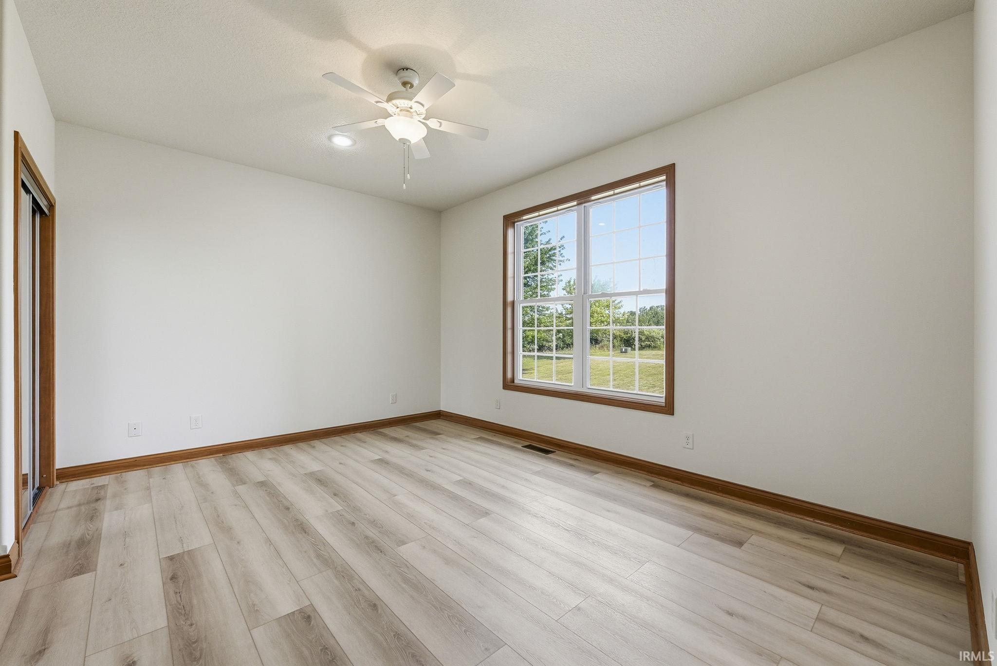 Spacious room featuring light wood-finish flooring, a large window with wood trim, a ceiling fan with integrated lighting, recessed ceiling light, and a sliding closet door with matching wood trim