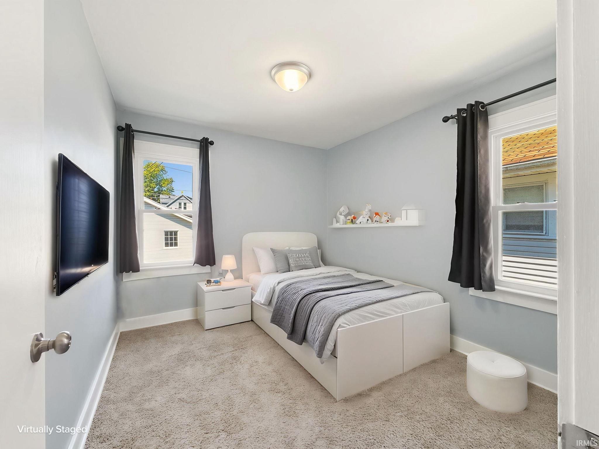 Light blue painted room featuring a wall-mounted television, two windows with dark curtains, a built-in wall shelf, and light-colored carpet flooring