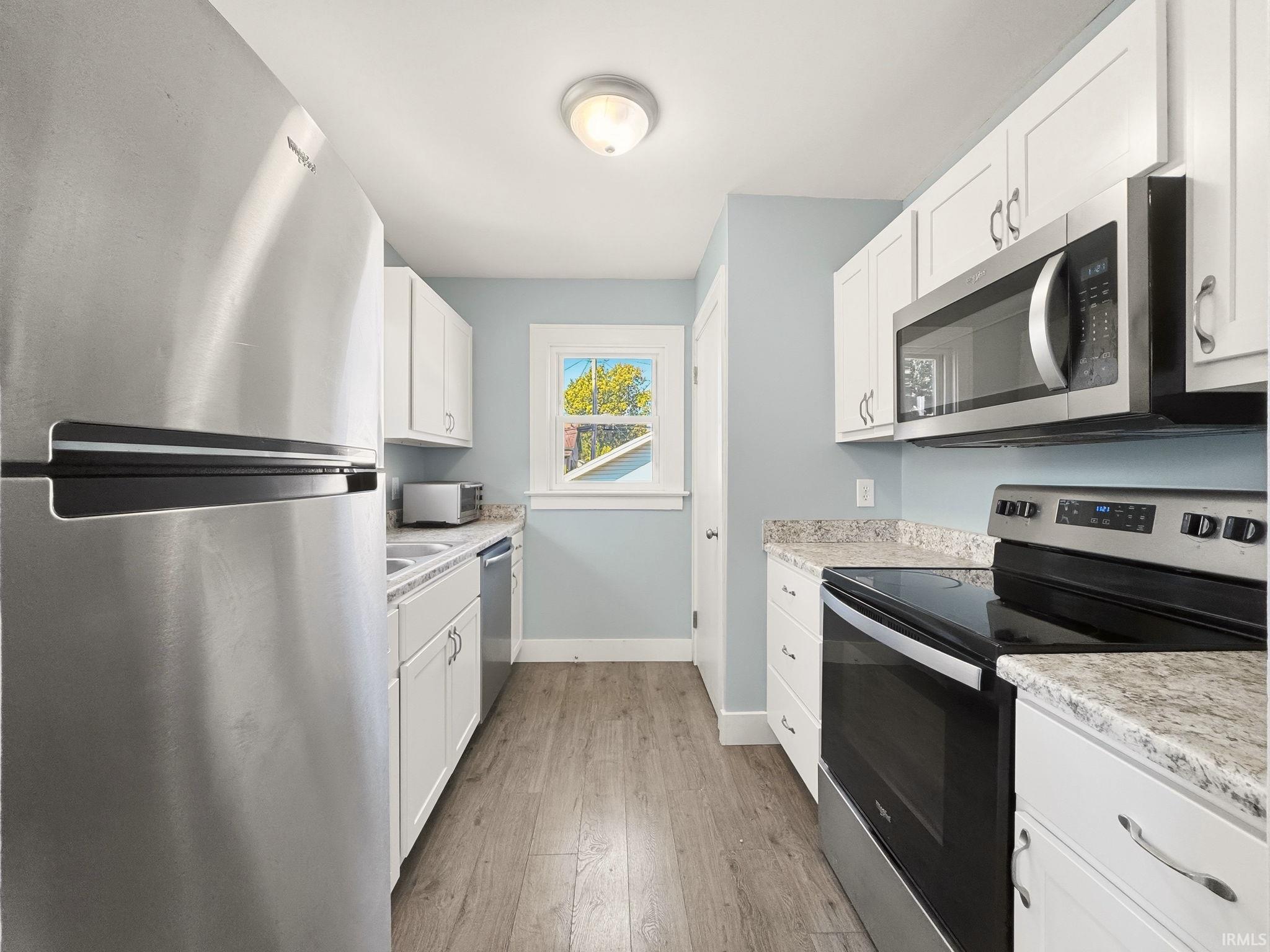 Modern kitchen featuring stainless steel appliances, white cabinetry, light-toned countertops, wood-finish flooring, and a window providing natural light