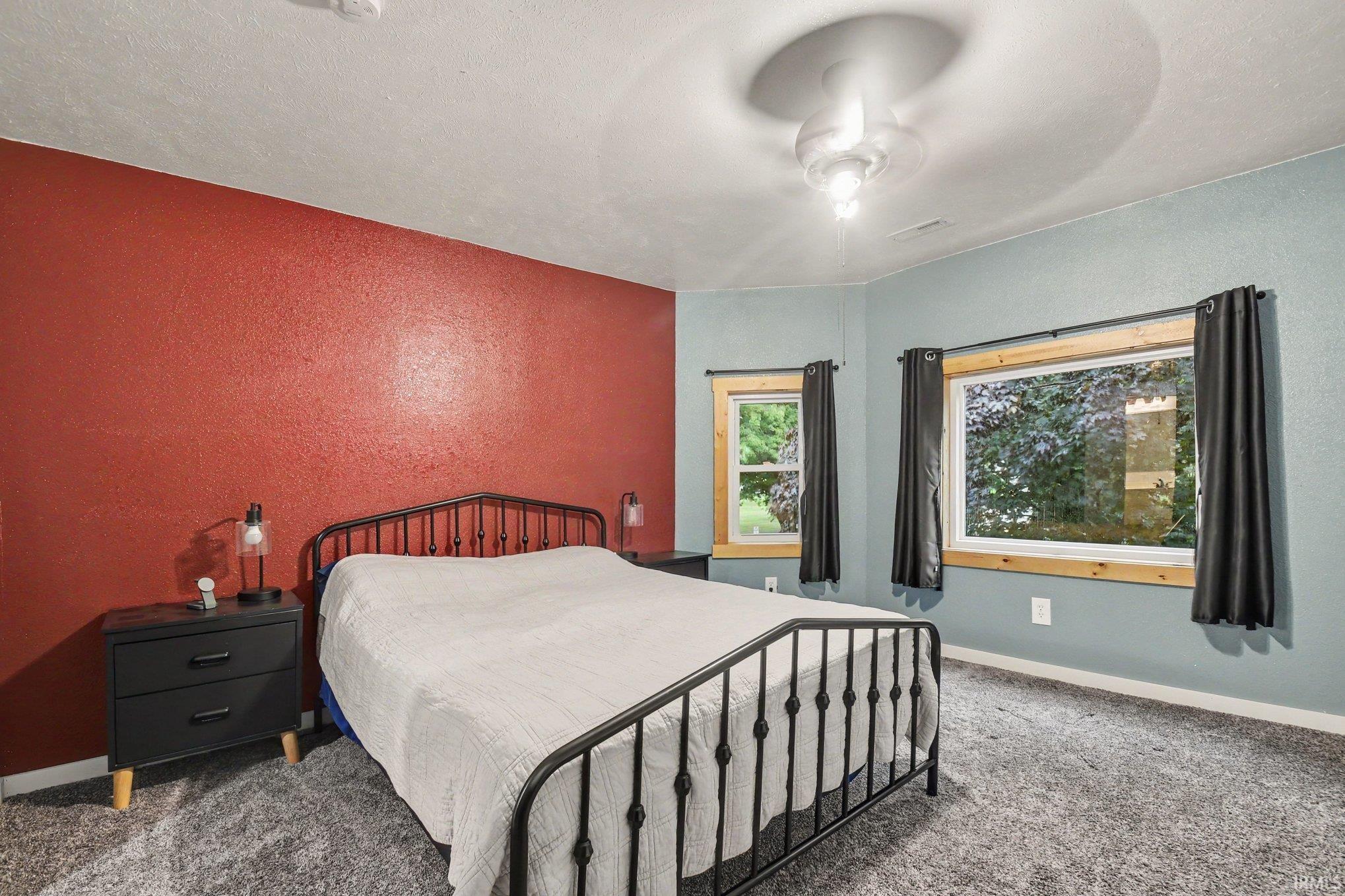 Bedroom featuring a textured accent wall, two windows with natural wood trim, and a ceiling fan with integrated lighting