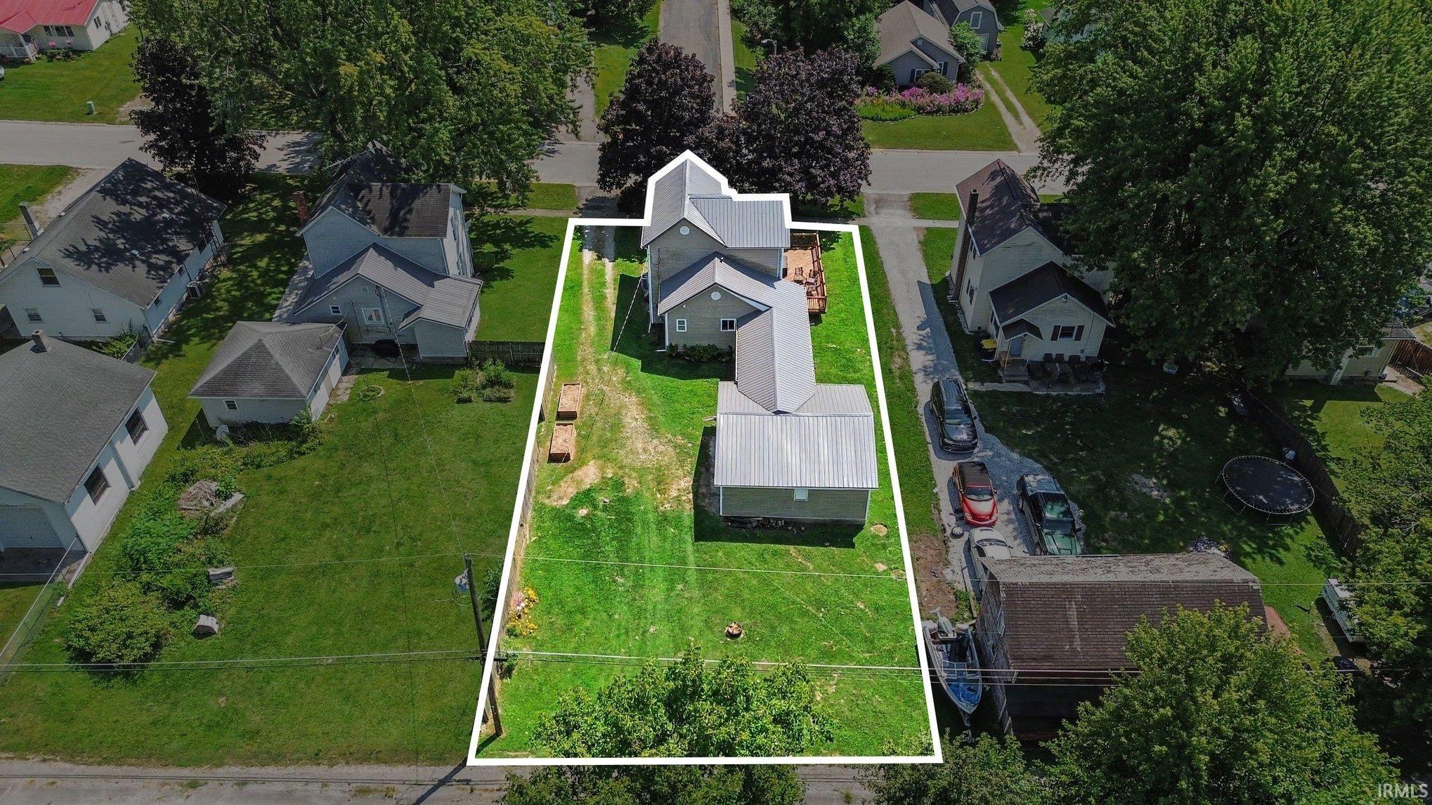 Aerial property depiction featuring a main residence, an elongated driveway, a detached outbuilding with a metal roof, a rear deck, and a grass-covered lot