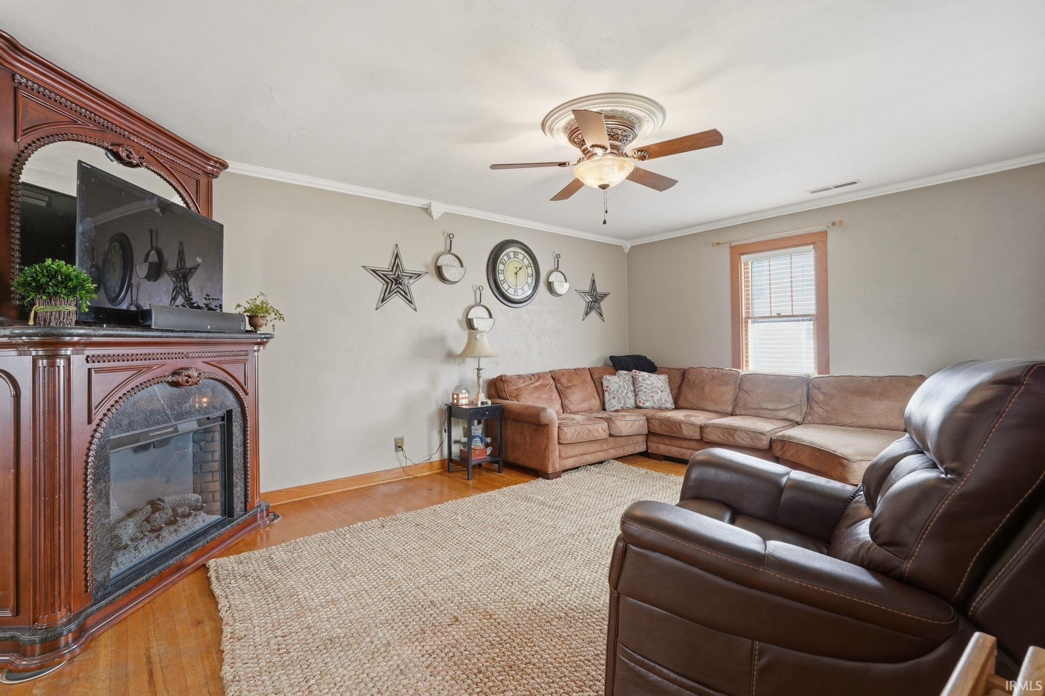Living area featuring wood-finish flooring, a decorative fireplace mantel with an electric insert, a ceiling fan with integrated lighting, and a single window with wood trim