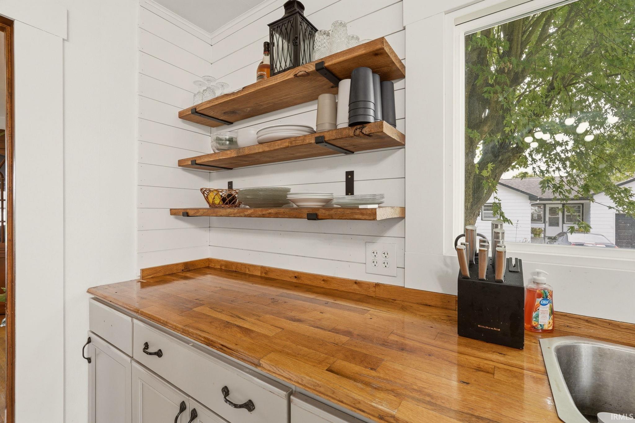 Chef's kitchen workspace showcasing warm wood-finish countertops, white shaker-style cabinetry with oil-rubbed bronze hardware, stainless steel undermount sink, and three floating wood shelves mounted above the counter