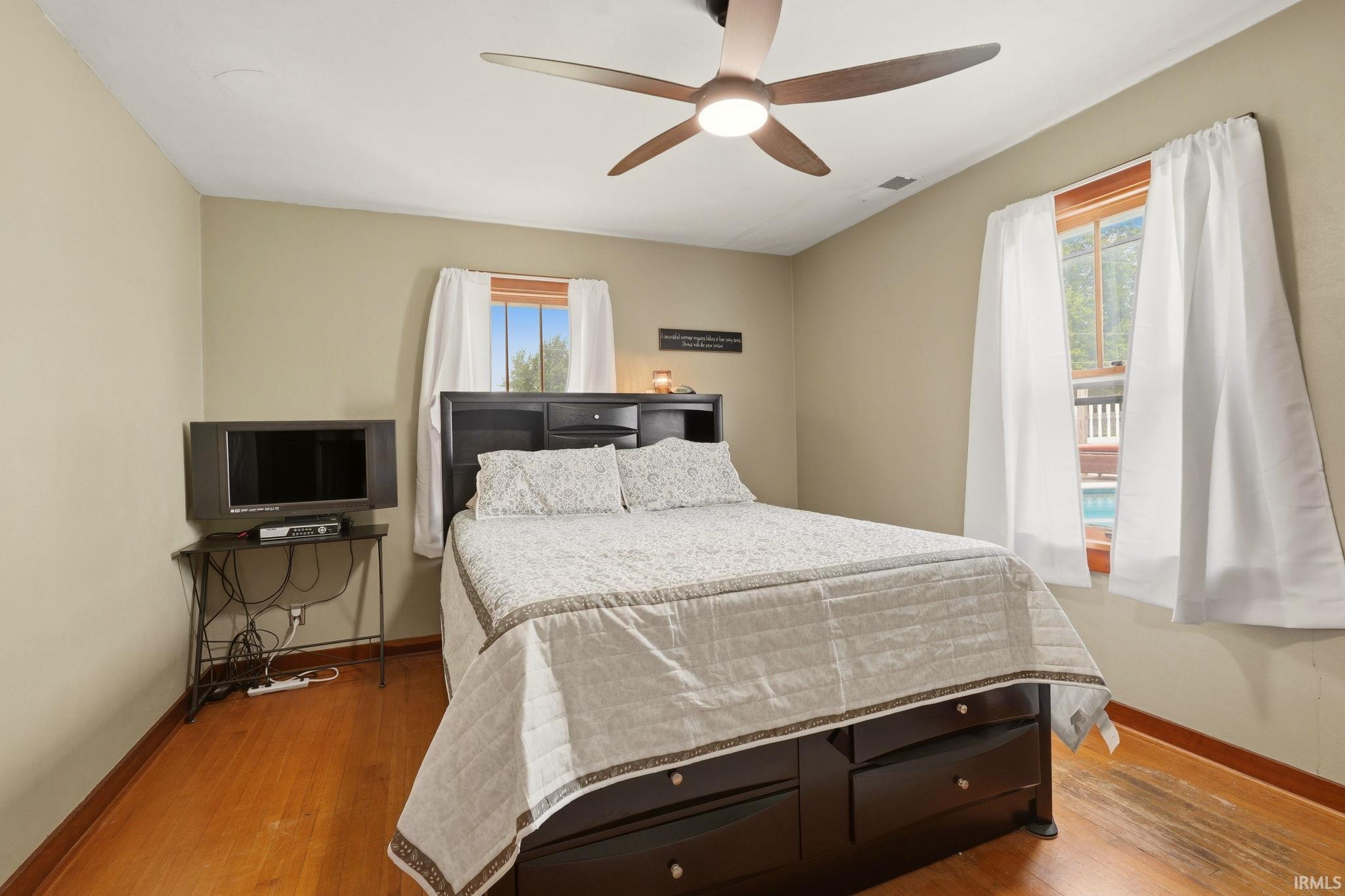 Bedroom featuring wood-finish flooring, two windows with wood frames, and a ceiling fan with integrated lighting