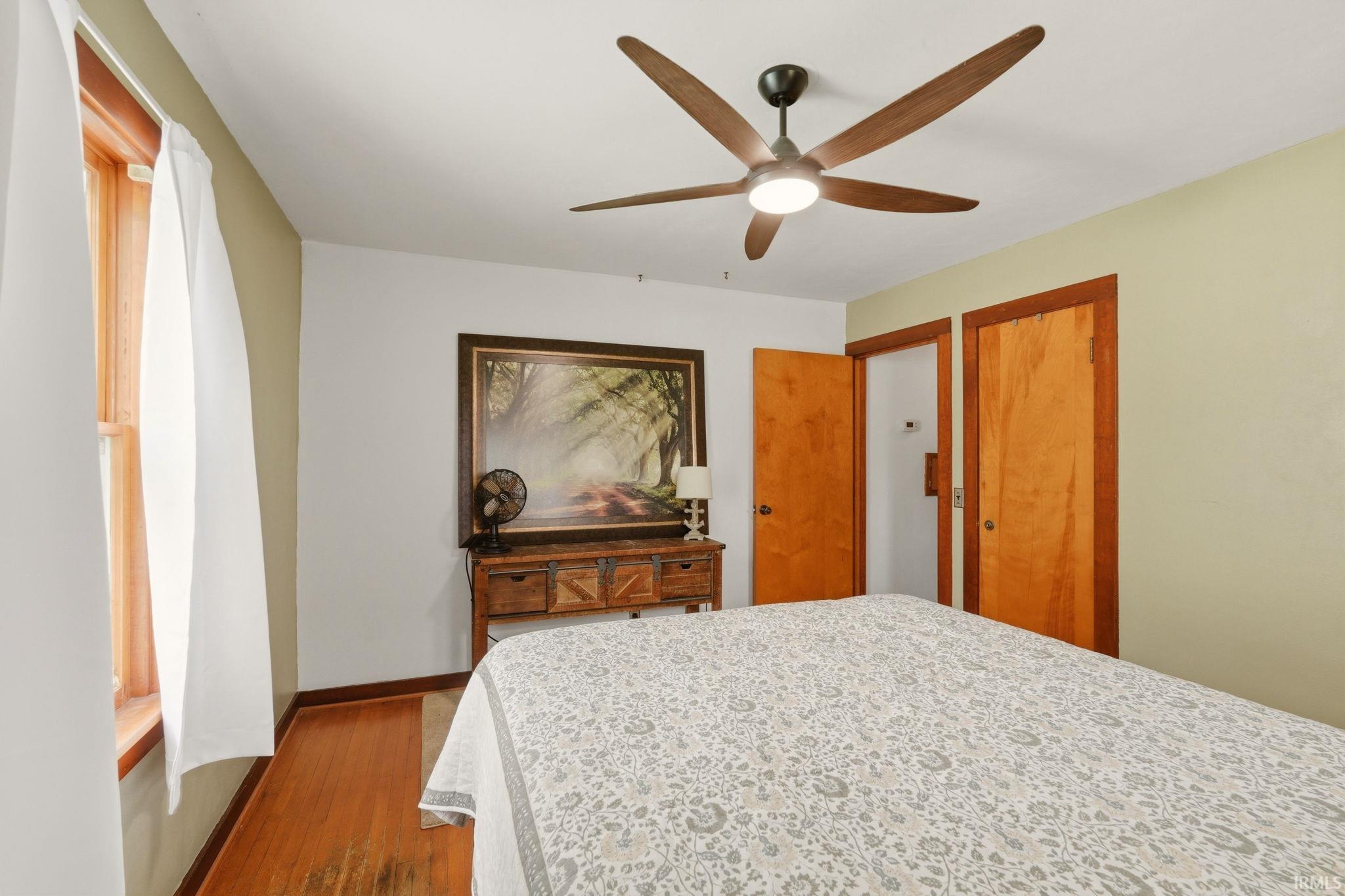Bedroom featuring wood-finish flooring, a ceiling fan with integrated lighting, and natural wood trim around the window and doors