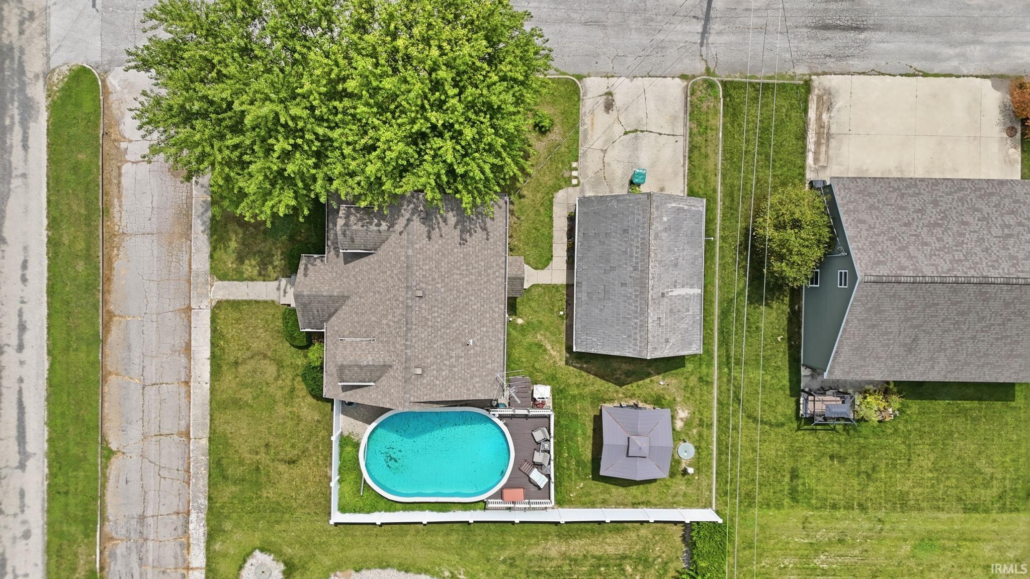 Aerial view of this residential property showcasing a sparkling in-ground swimming pool with surrounding concrete decking, manicured lawn areas, mature shade trees, attached garage, and detached outbuilding