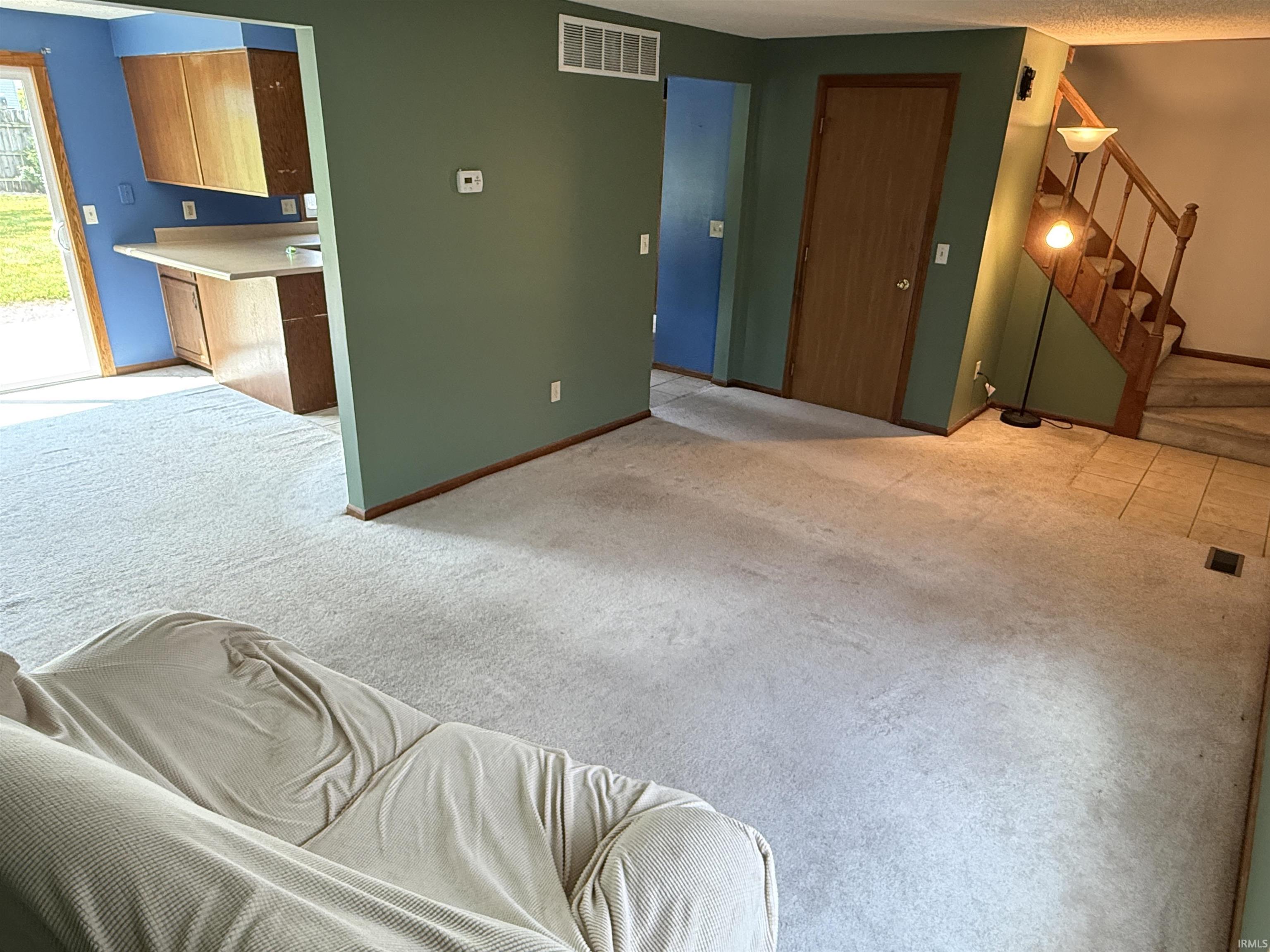 Open concept living area with carpeting, featuring a kitchen pass-through, a full staircase with wood banister and steps, and a sliding glass door to exterior