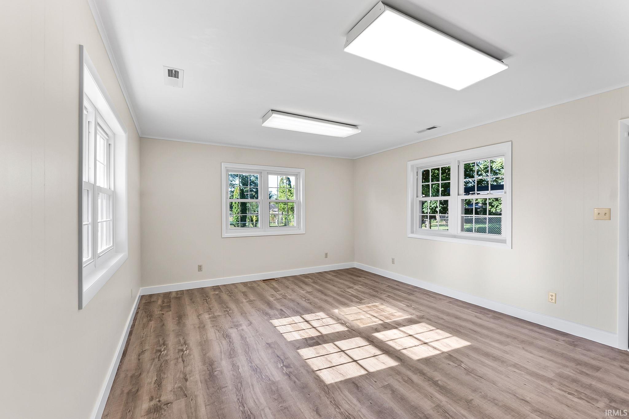Bright, naturally lit interior space featuring wood-finish flooring, multiple casement windows with grid detailing, recessed ceiling lighting, and pristine white walls with electrical outlets throughout