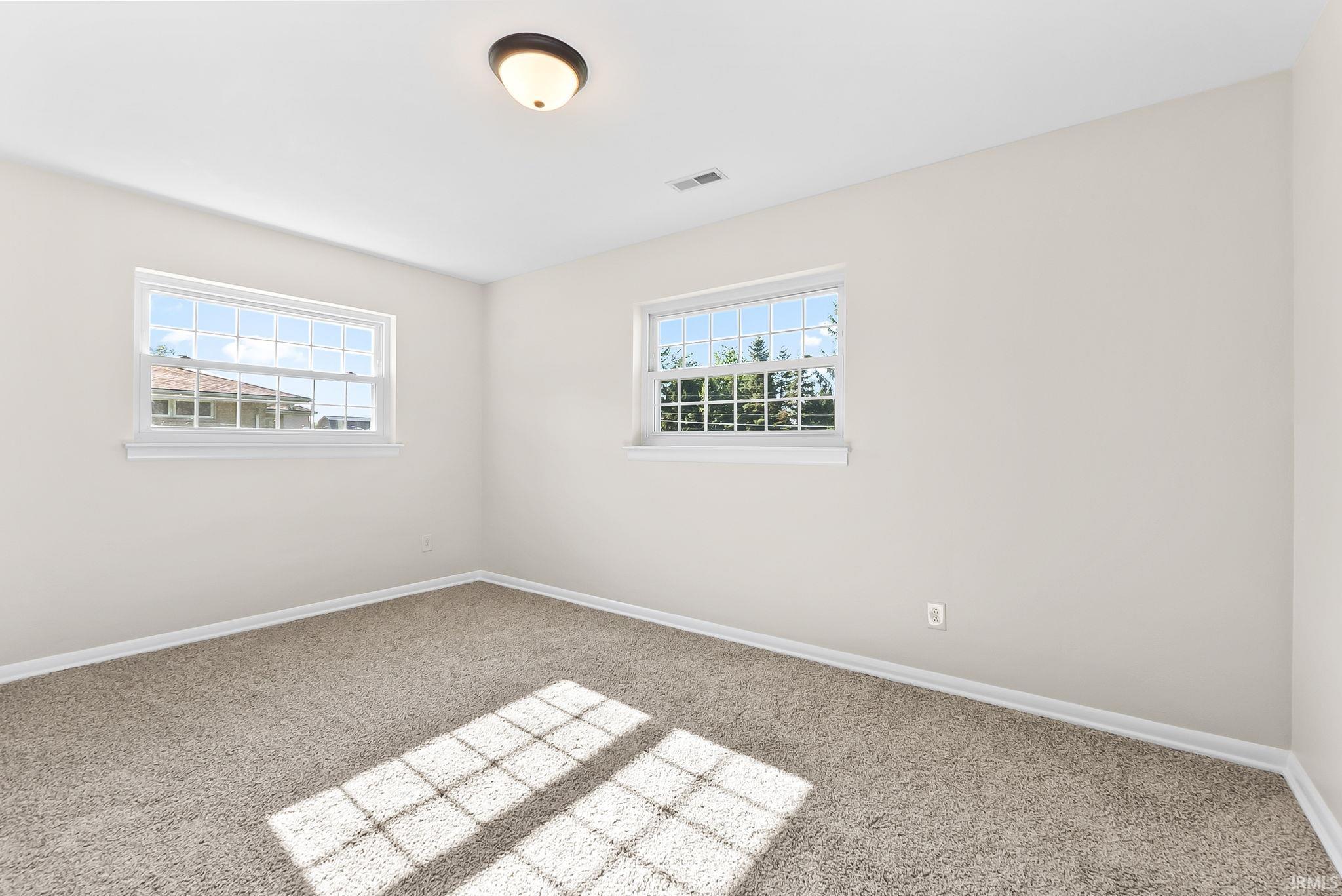 Bright secondary bedroom flooded with natural light from dual windows, featuring neutral wall finishes, neutral-toned carpet flooring, and a ceiling-mounted light fixture