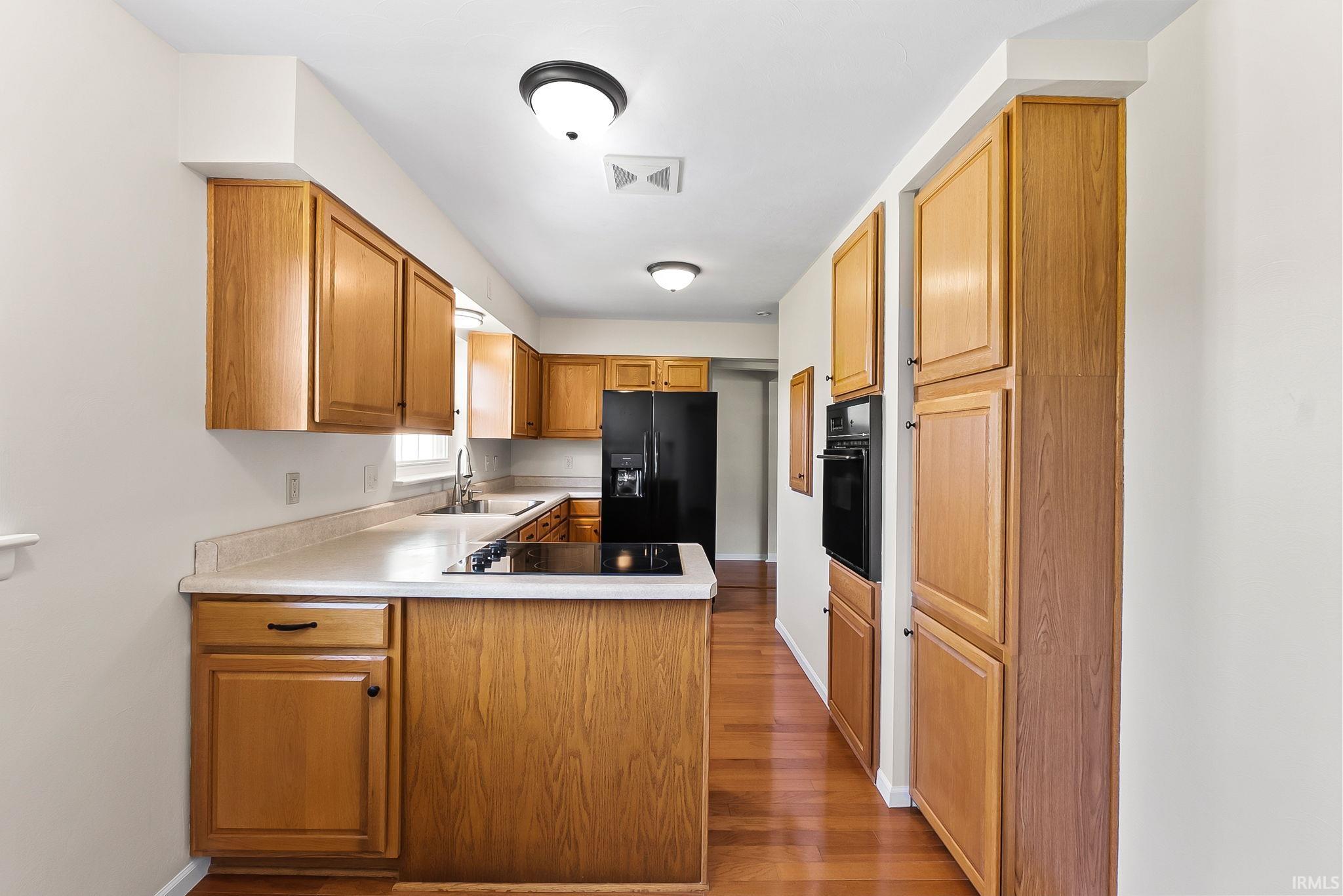 Galley kitchen featuring wood-finish cabinetry, light-toned countertops, a black electric cooktop, and a built-in black wall oven