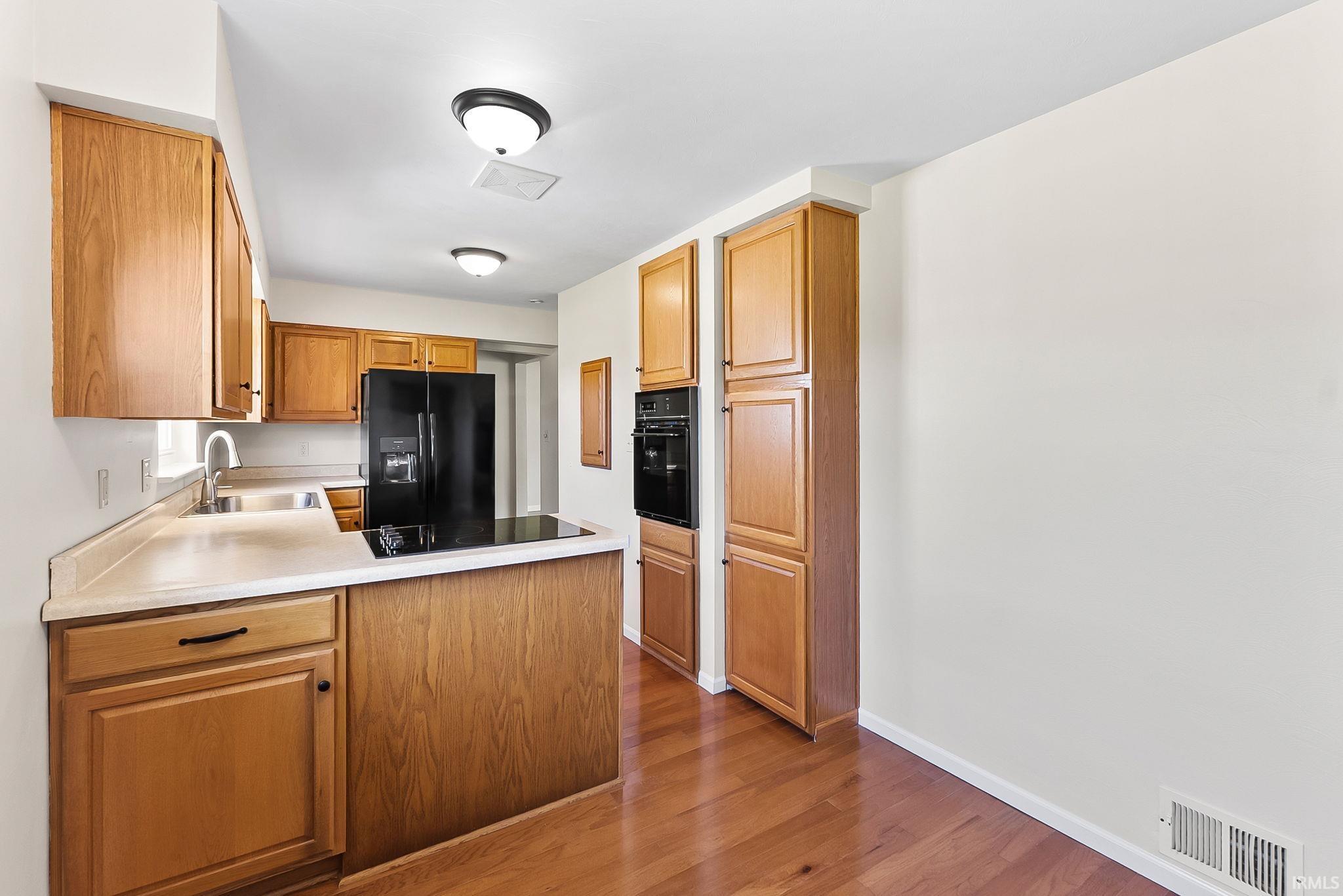 Kitchen featuring wood-finish cabinetry, light-toned countertops, a black refrigerator, built-in oven, and a cooktop