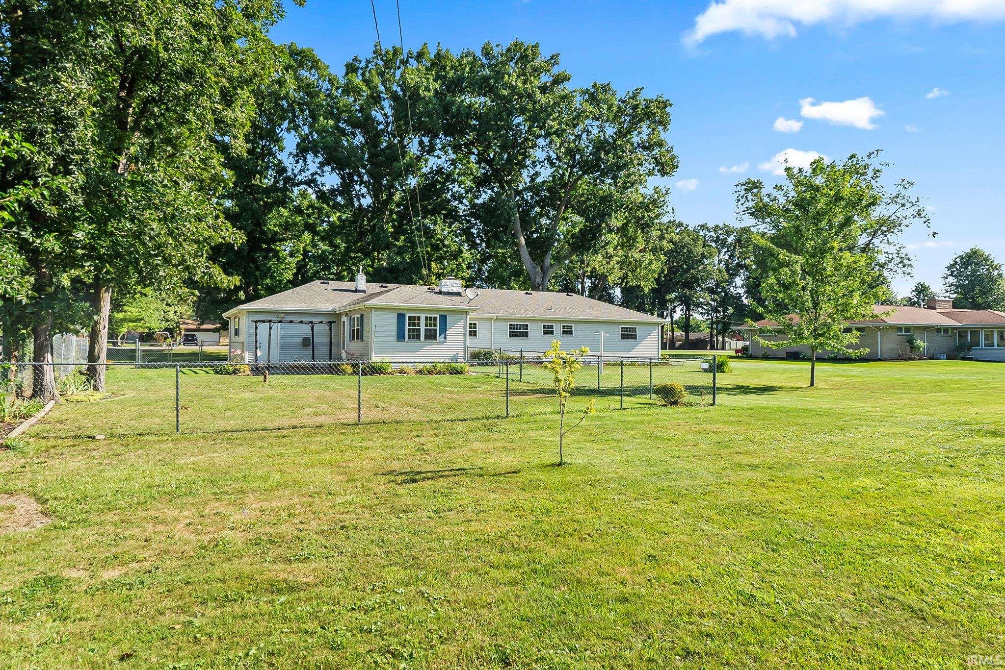 Expansive grounds featuring a manicured lawn bordered by mature shade trees and fencing, with a single-story ranch-style residence offering a covered front porch and multiple windows for natural light