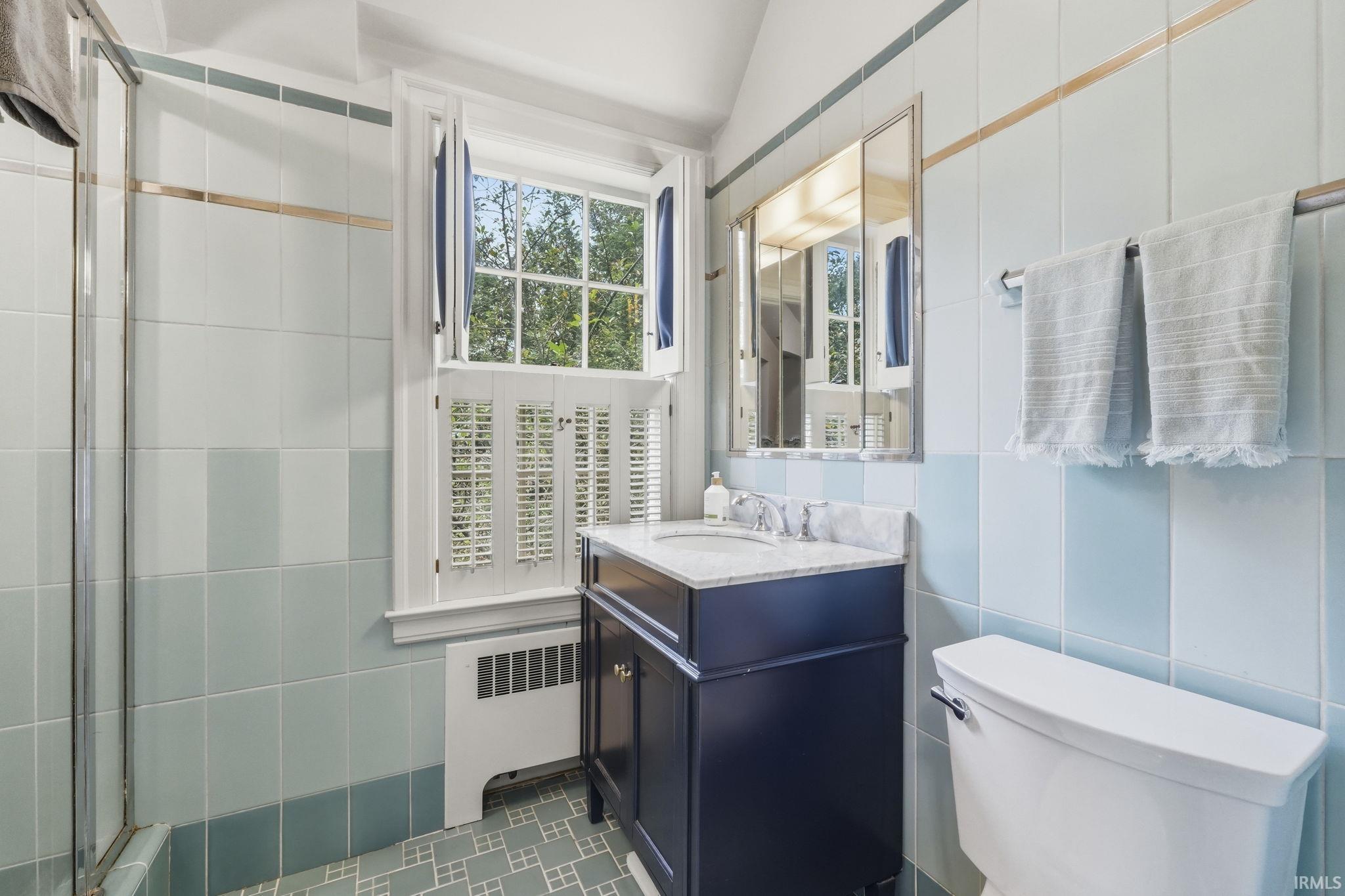 Light blue and white tiled bathroom with a window featuring white plantation shutters, a dark wood vanity with a white basin and stone countertop, and a mirrored medicine cabinet