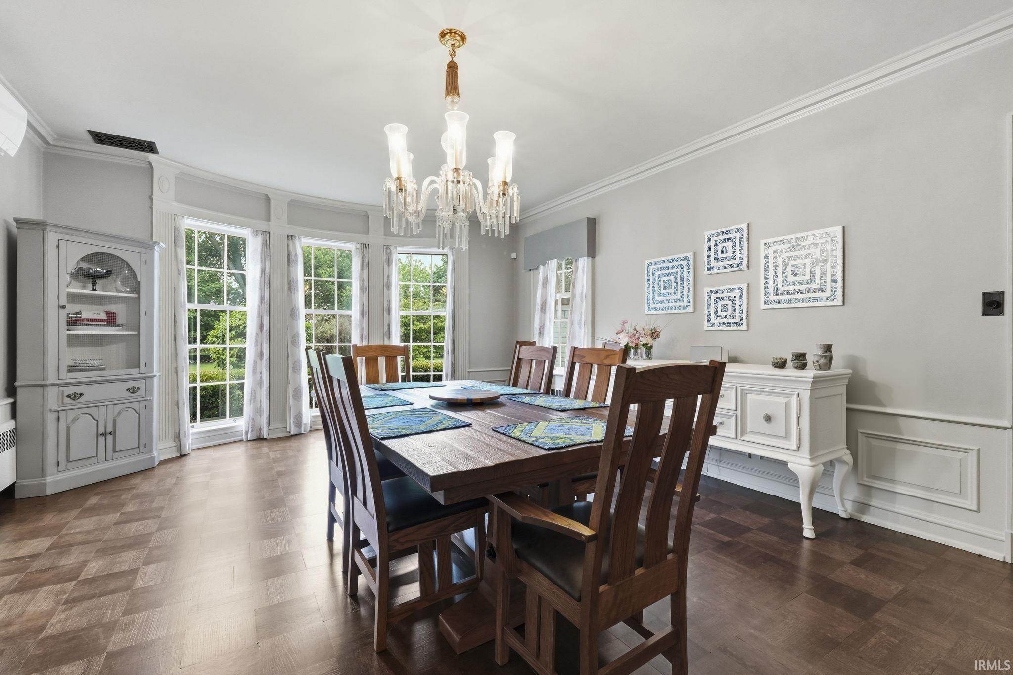 Chandelier lighting with crystal accents, wood-finish flooring with a parquet pattern, curved window wall with multiple window panes, crown molding, and wainscoting details