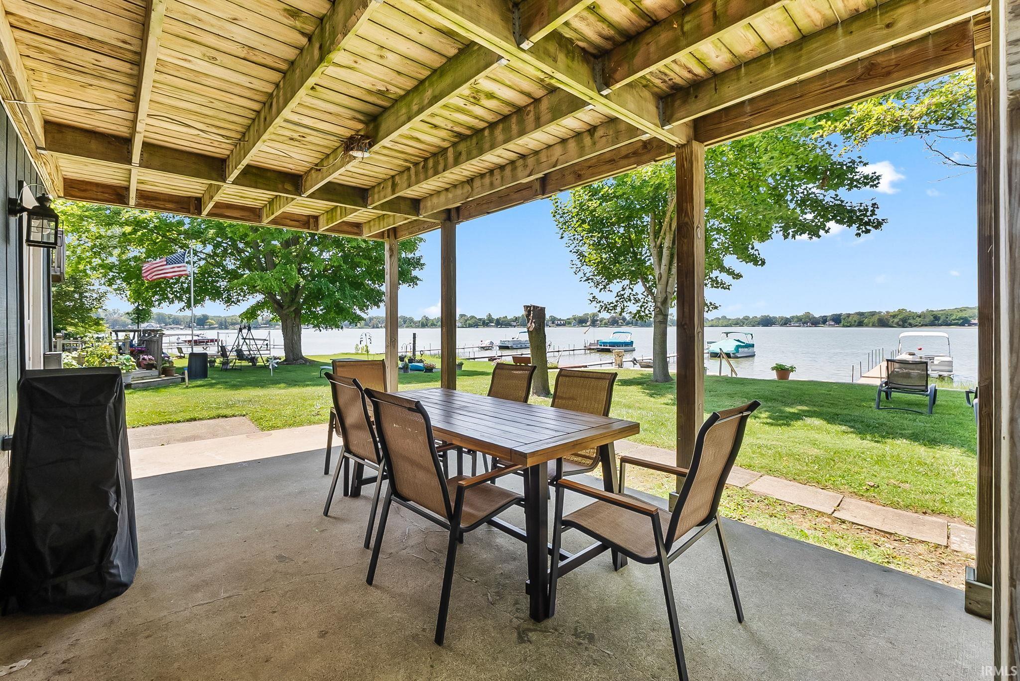 Covered concrete patio with exposed wood ceiling beams, adjacent to a manicured lawn, and expansive waterfront access