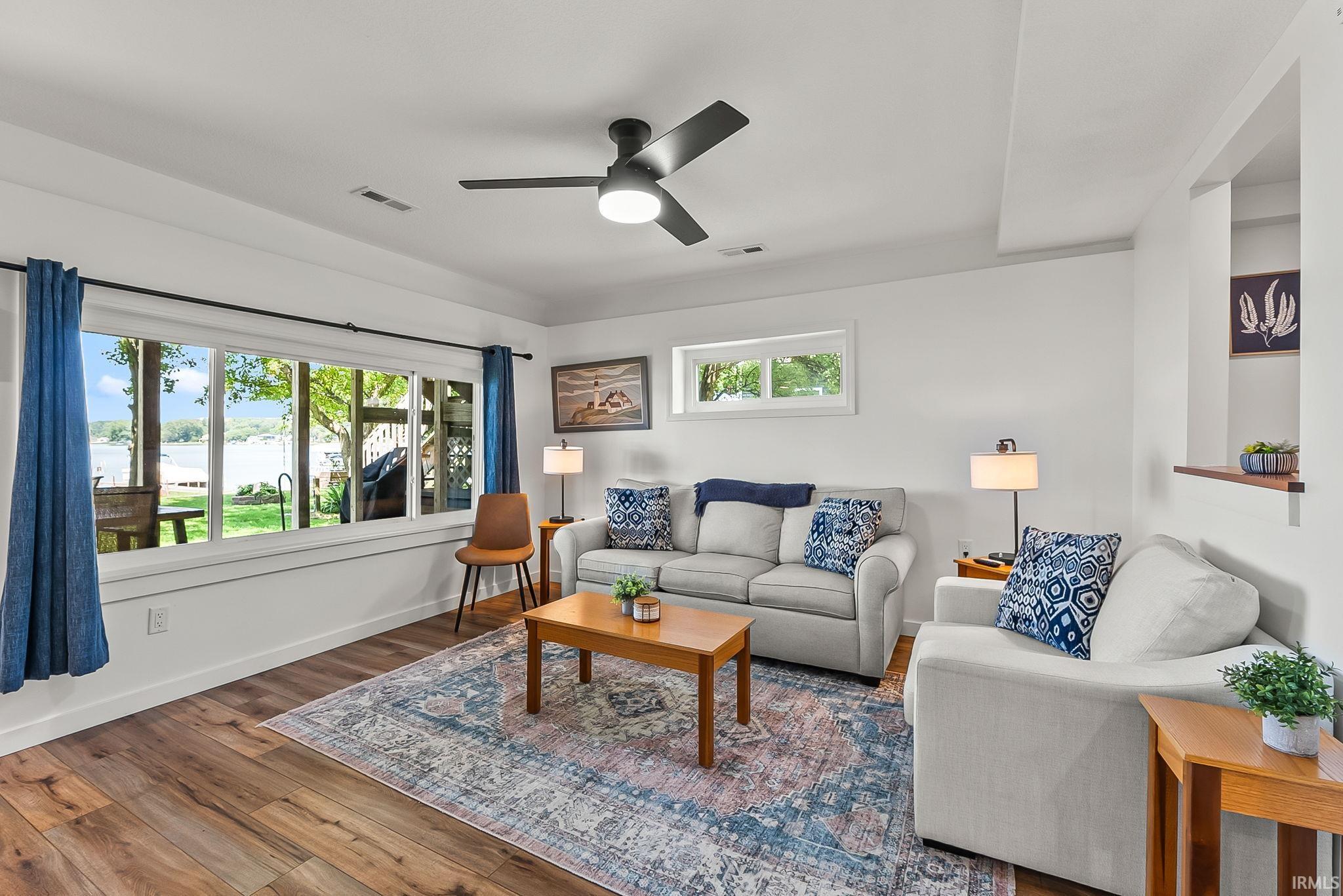 Bright, open living space showcasing wood-finish flooring, abundant natural light from multi-pane glass doors and windows, contemporary ceiling fan with integrated lighting, and neutral upholstered seating arrangements