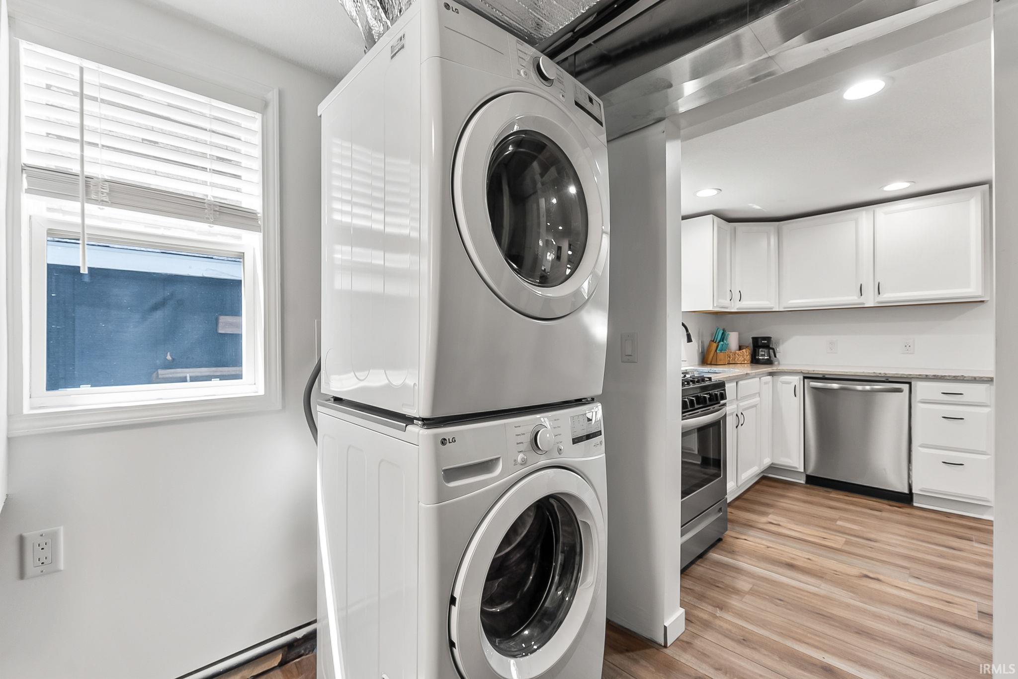 Modern laundry room featuring stacked front-load washers and dryers with white cabinetry, stainless steel appliances, and wood-finish flooring