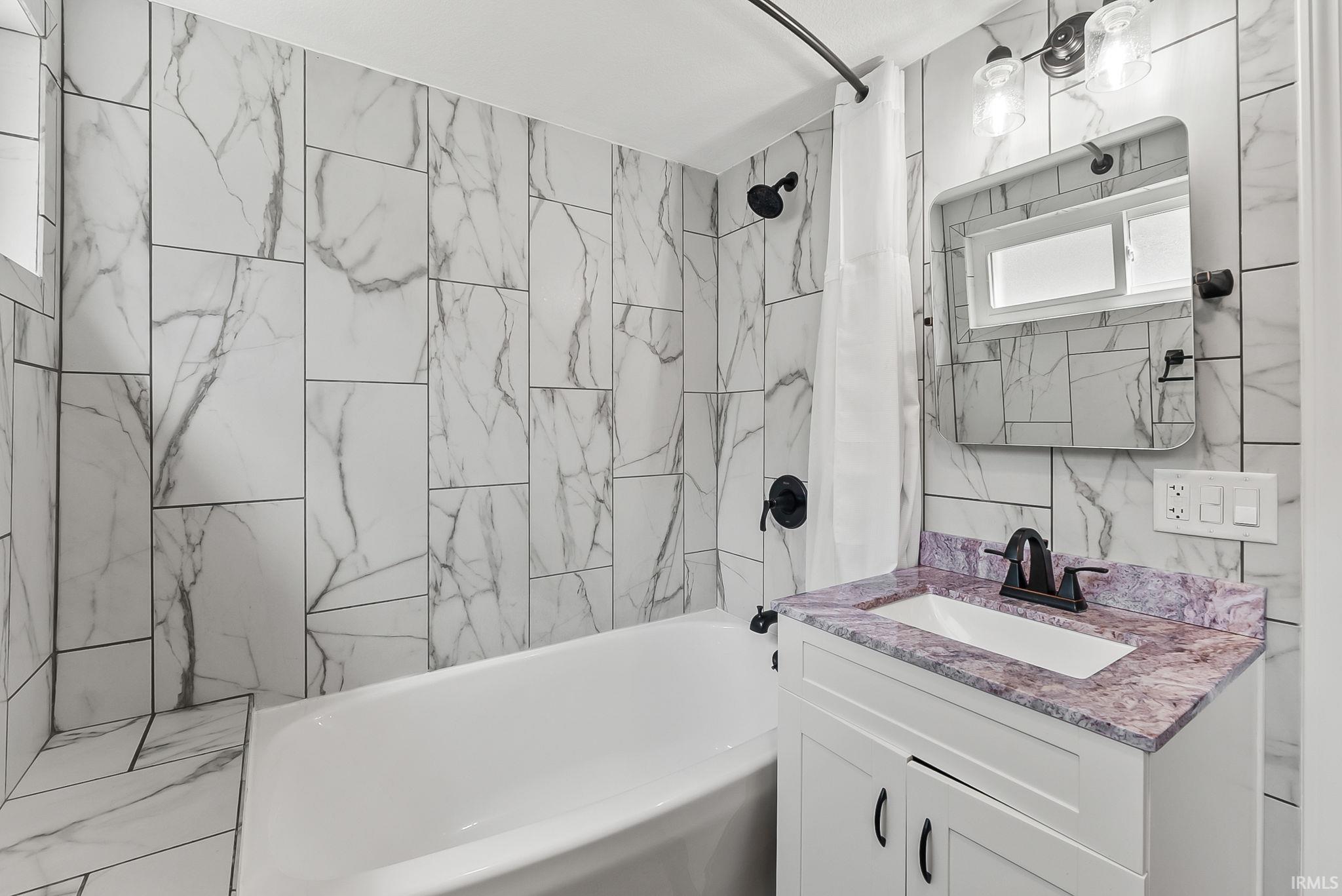 Bathtub with a shower, patterned tile surround, white vanity with a rectangular sink, purple-toned countertop, and matte black fixtures