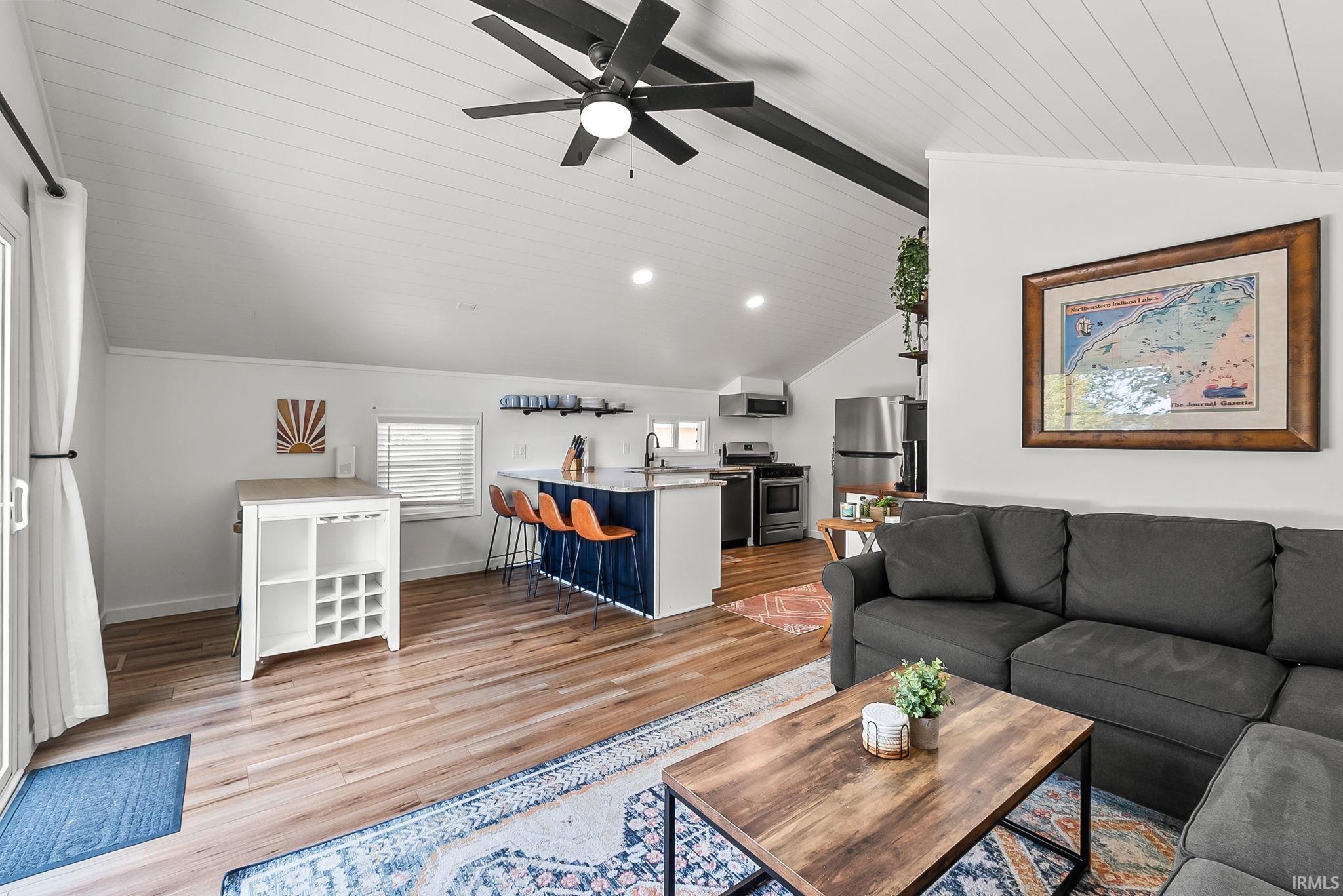 Vaulted ceiling with exposed beams, wood-finish flooring, kitchen island with seating, stainless steel appliances, and built-in shelving
