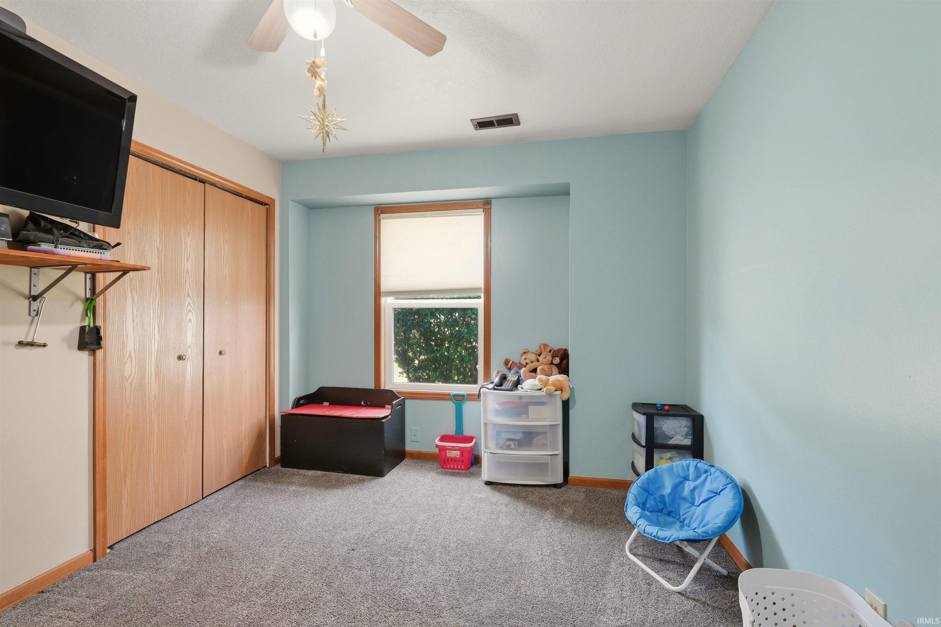 Carpeted room with a ceiling fan, a window with a roller shade, and built-in closet doors with wood-finish panels