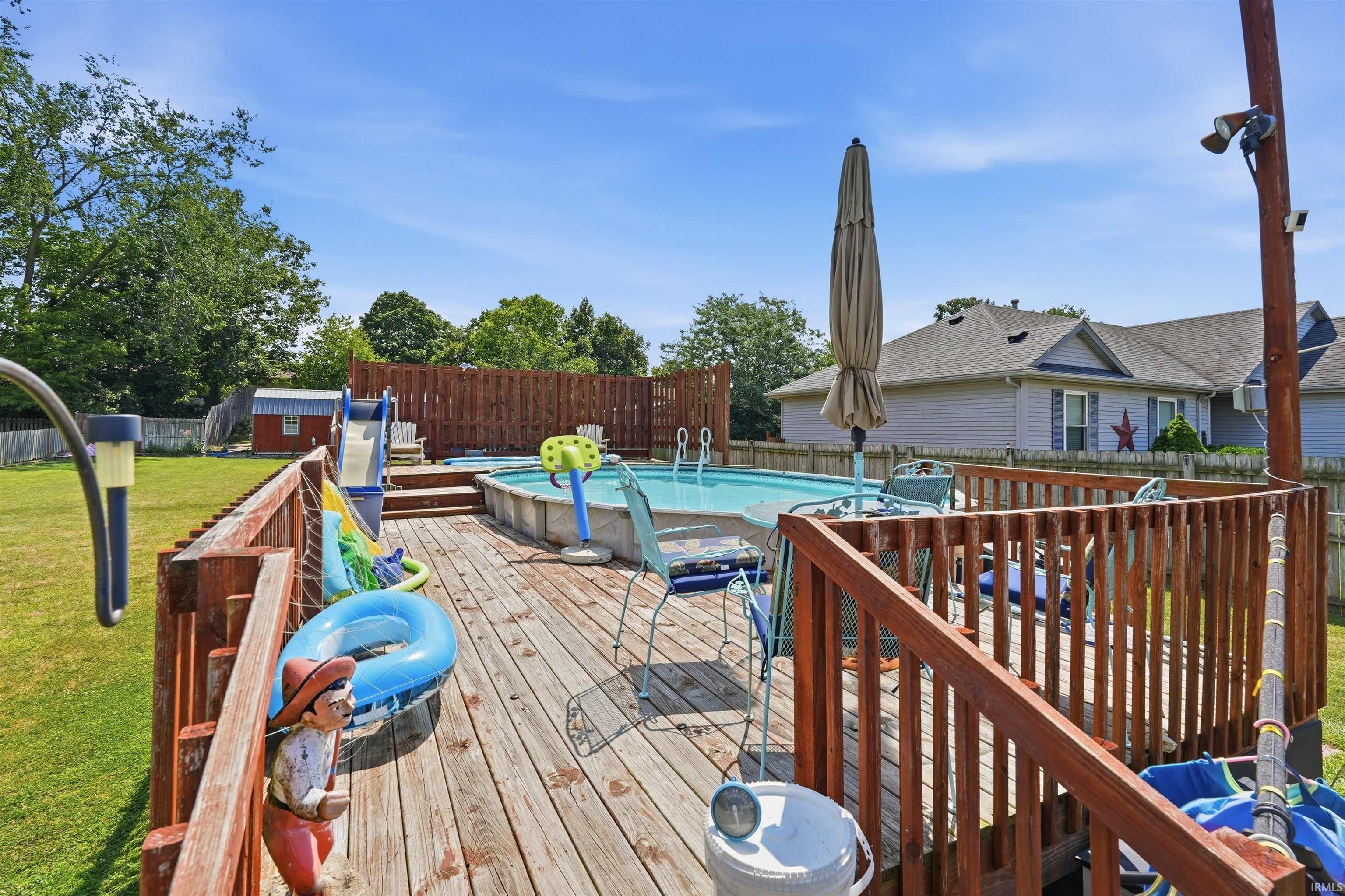 Expansive wood deck with railing surrounds an above-ground pool, featuring a small storage shed and mature trees