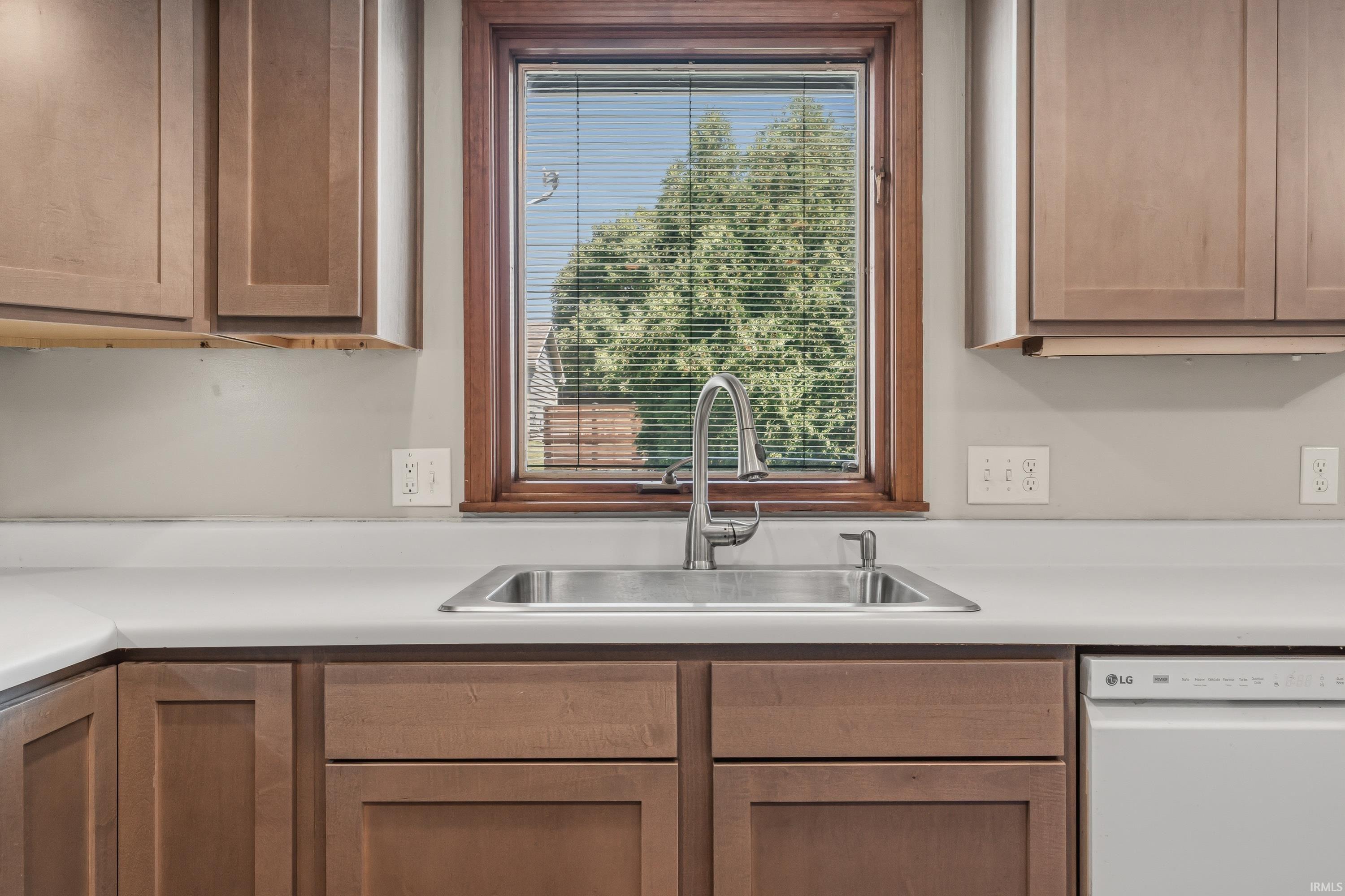 Stainless steel sink with a gooseneck faucet, light-toned countertops, wood-finish cabinetry, and a white dishwasher