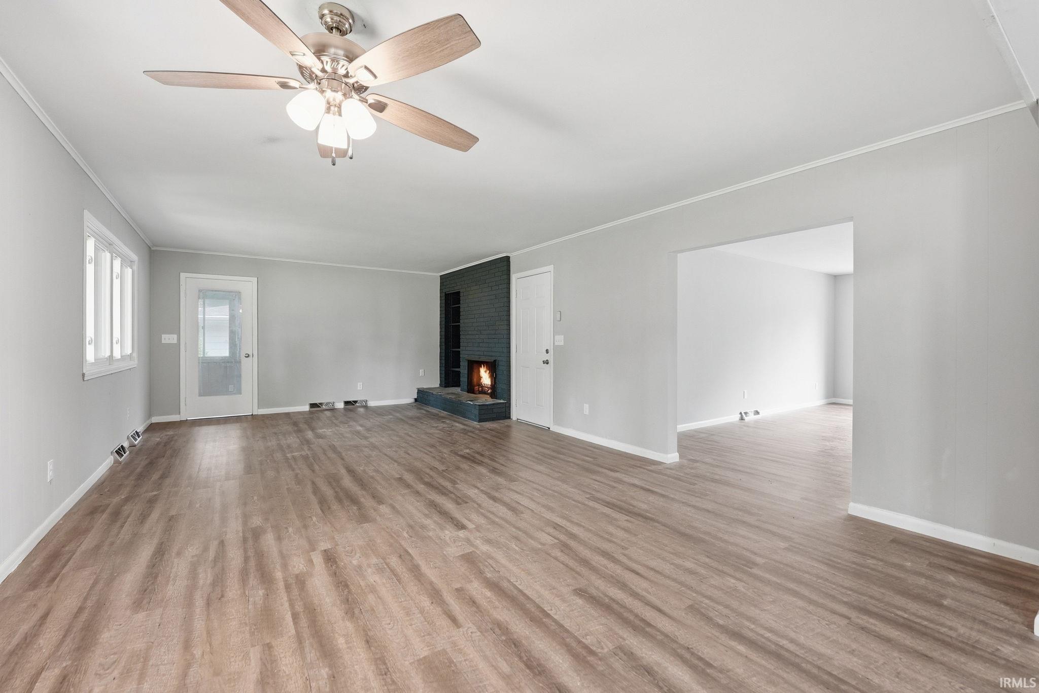 Spacious room featuring wood-finish flooring, a ceiling fan with integrated lighting, multiple windows, a fireplace with a dark brick surround, and an open entryway to an additional room